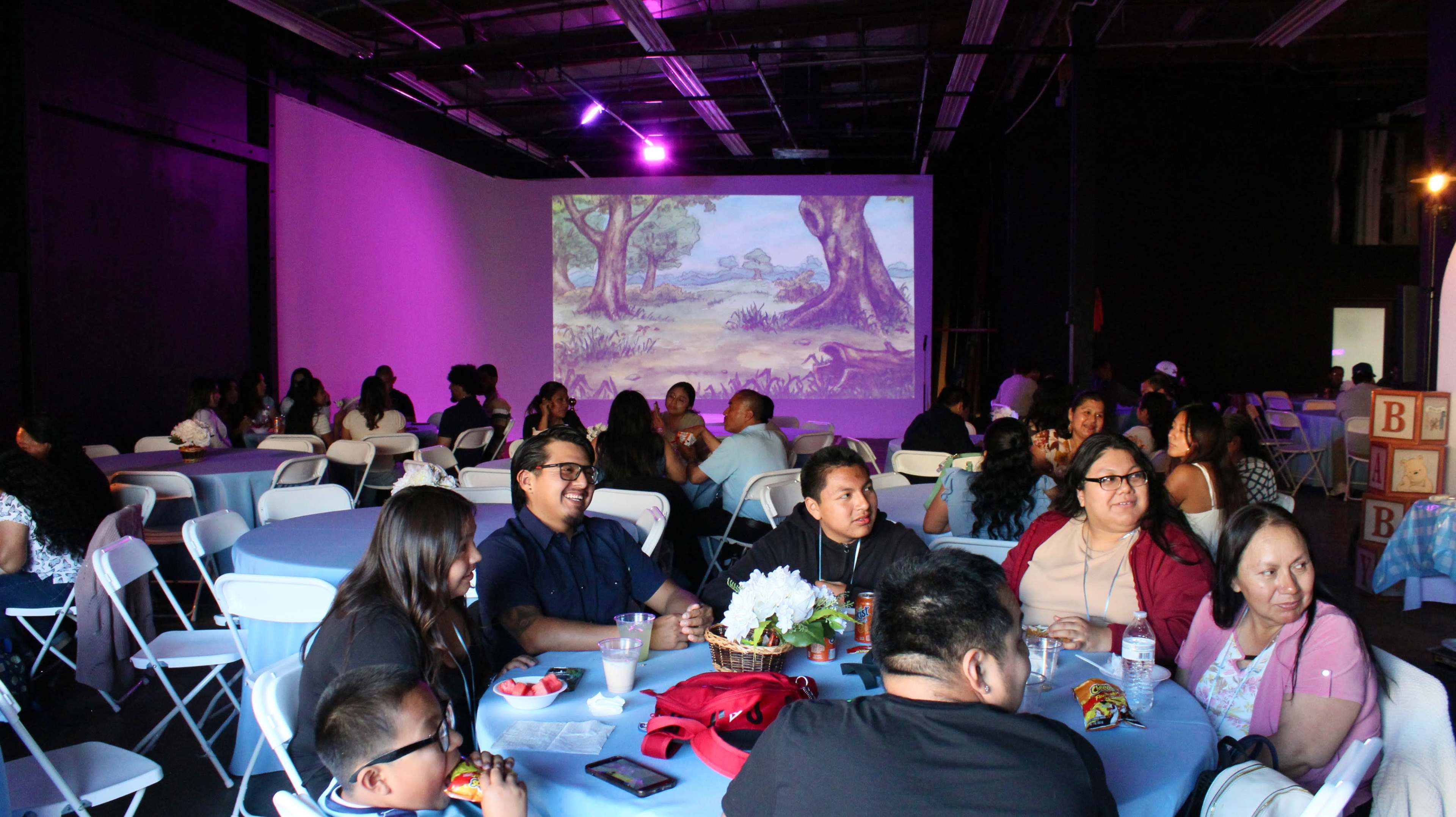 A large group of people sits at tables with blue tablecloths in a dimly lit event space featuring a projected image of trees on a wall.