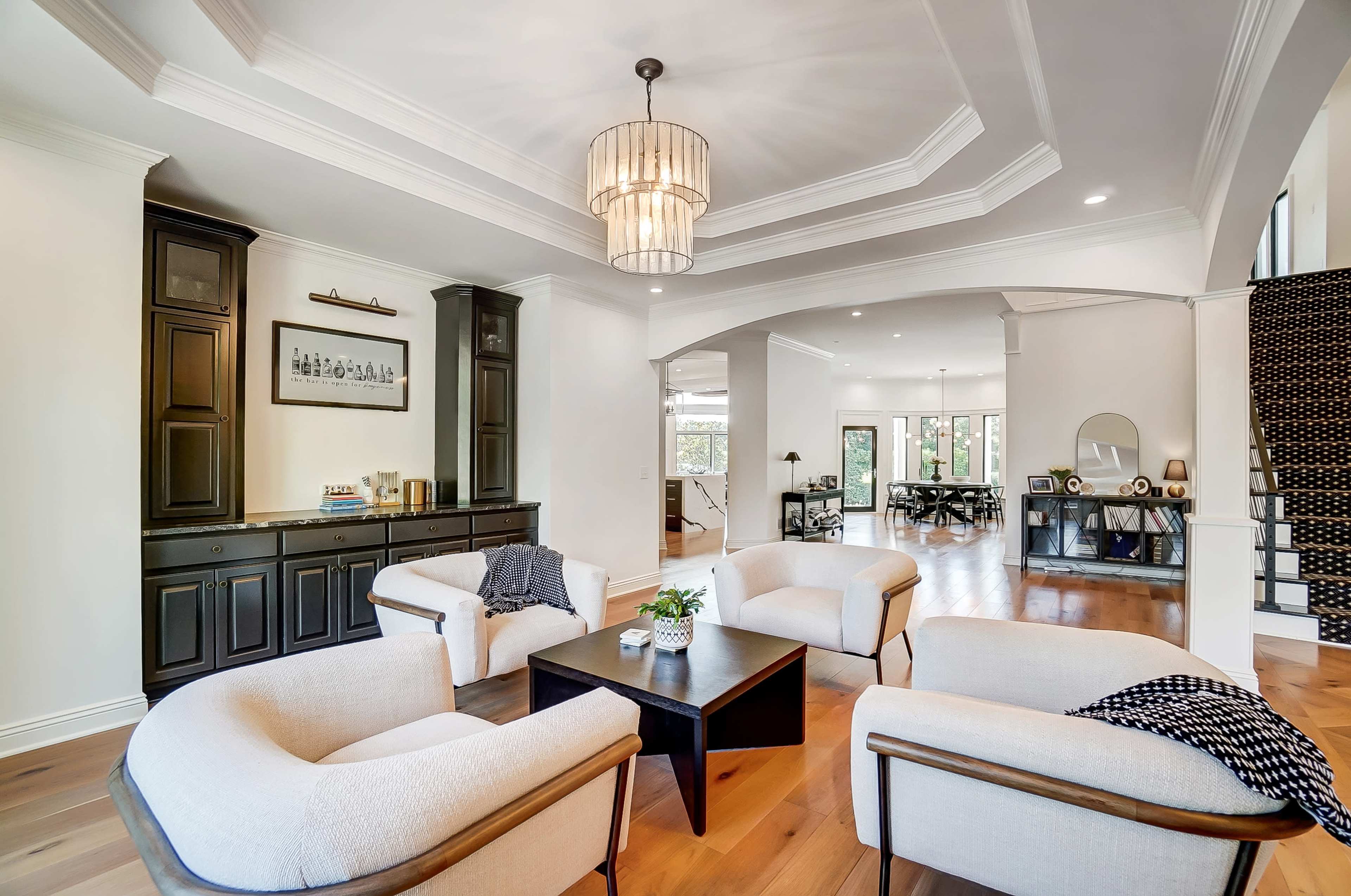 A spacious living area furnished with two armchairs, a coffee table, and a dark wooden cabinet, with an open view leading to another room and a staircase in the background.
