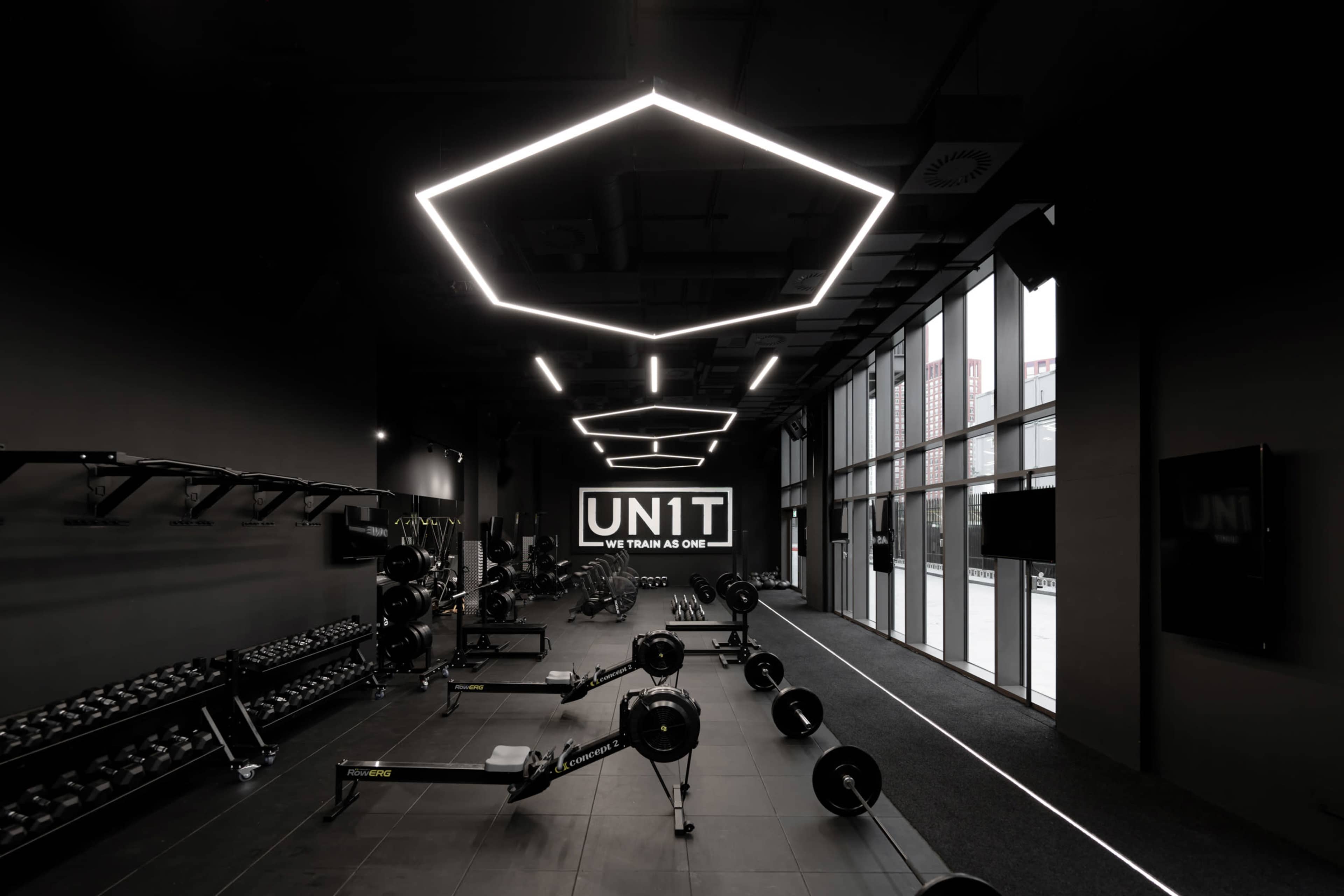 A modern gym interior featuring rows of weights and rowing machines under geometric LED lighting.