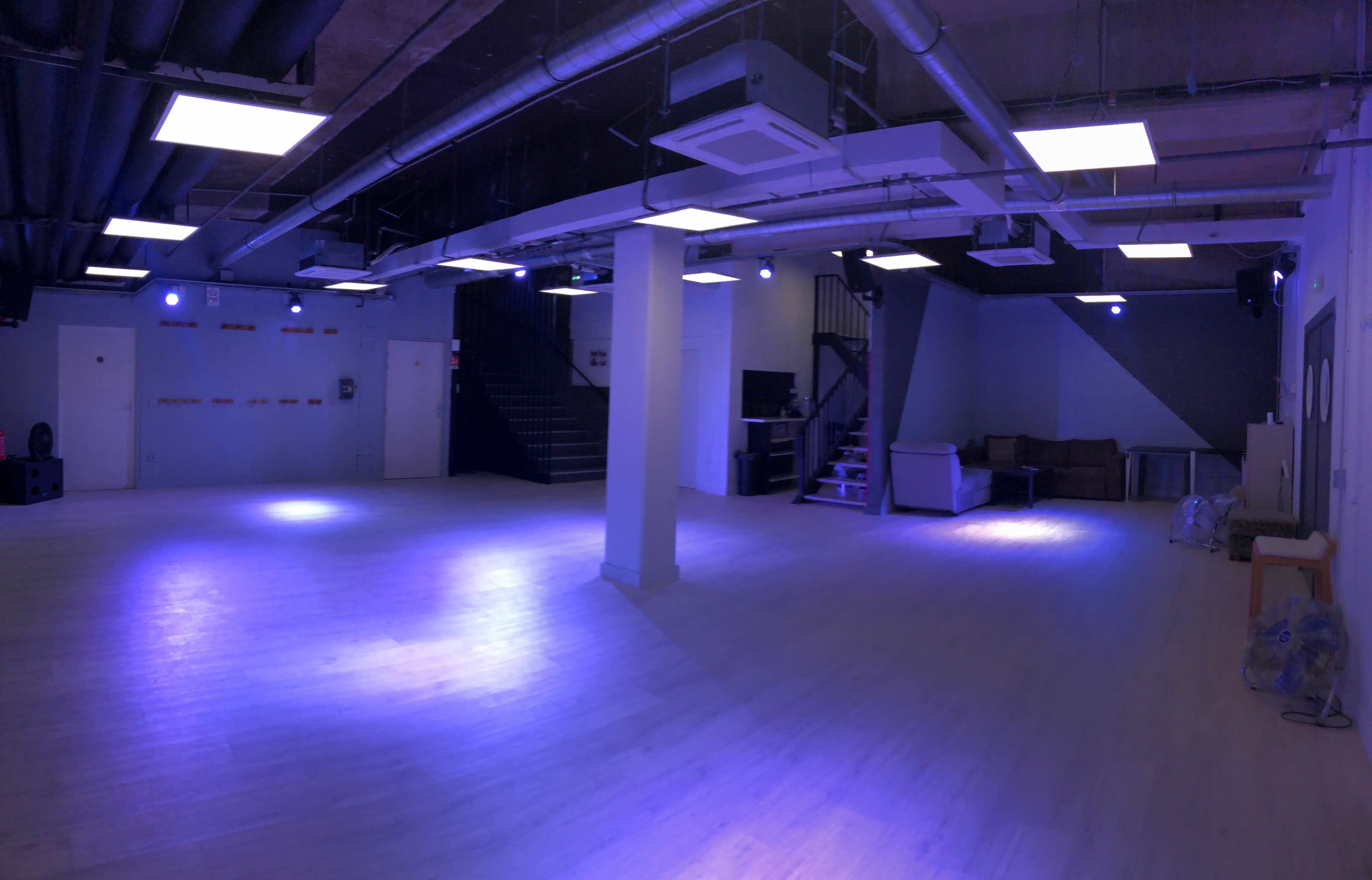 A spacious, dimly lit room with a light blue hue, featuring a staircase at one end and various seating arrangements along the walls.