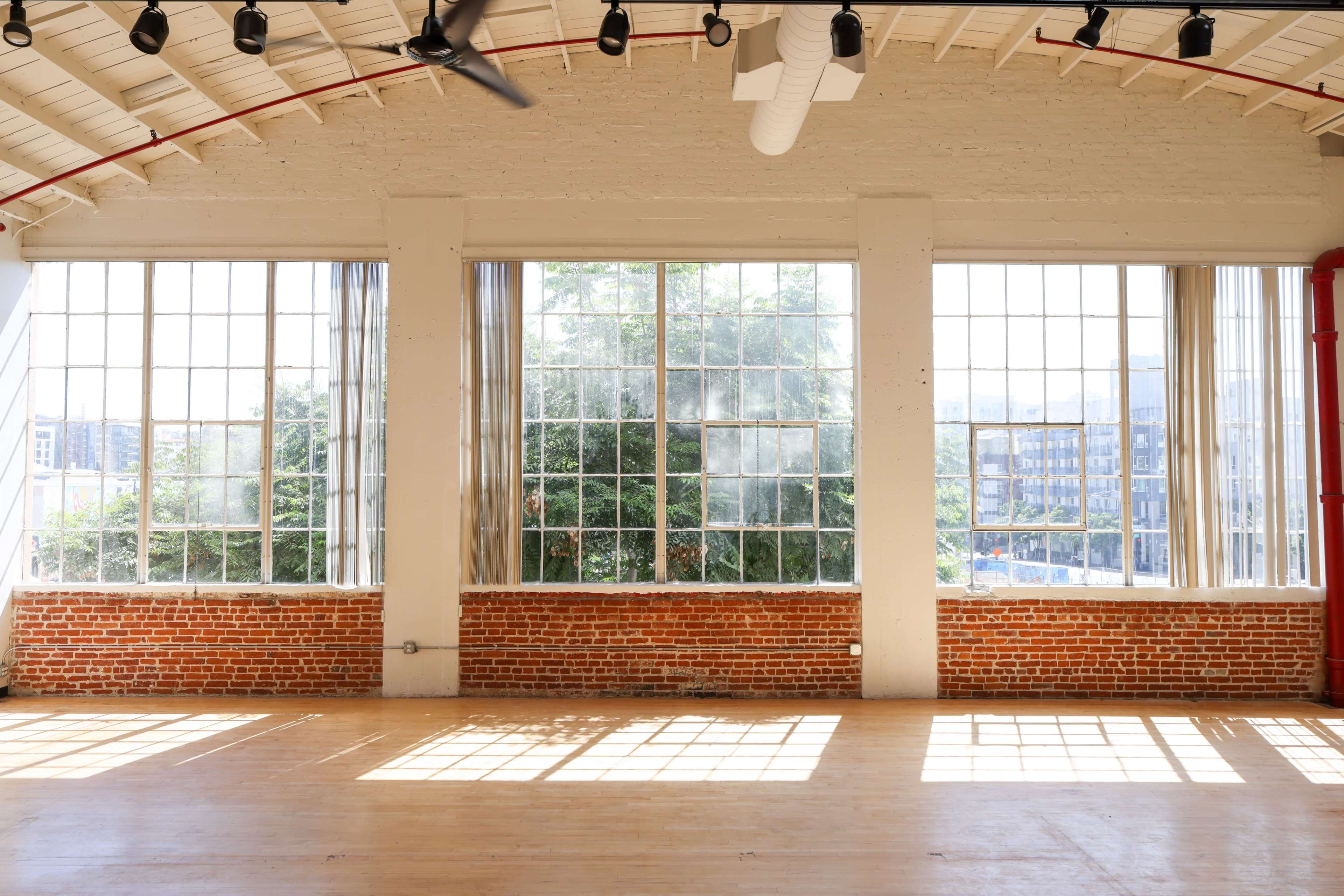 The image shows a spacious room with large windows that allow natural light to illuminate the wooden floor and reveal greenery outside.