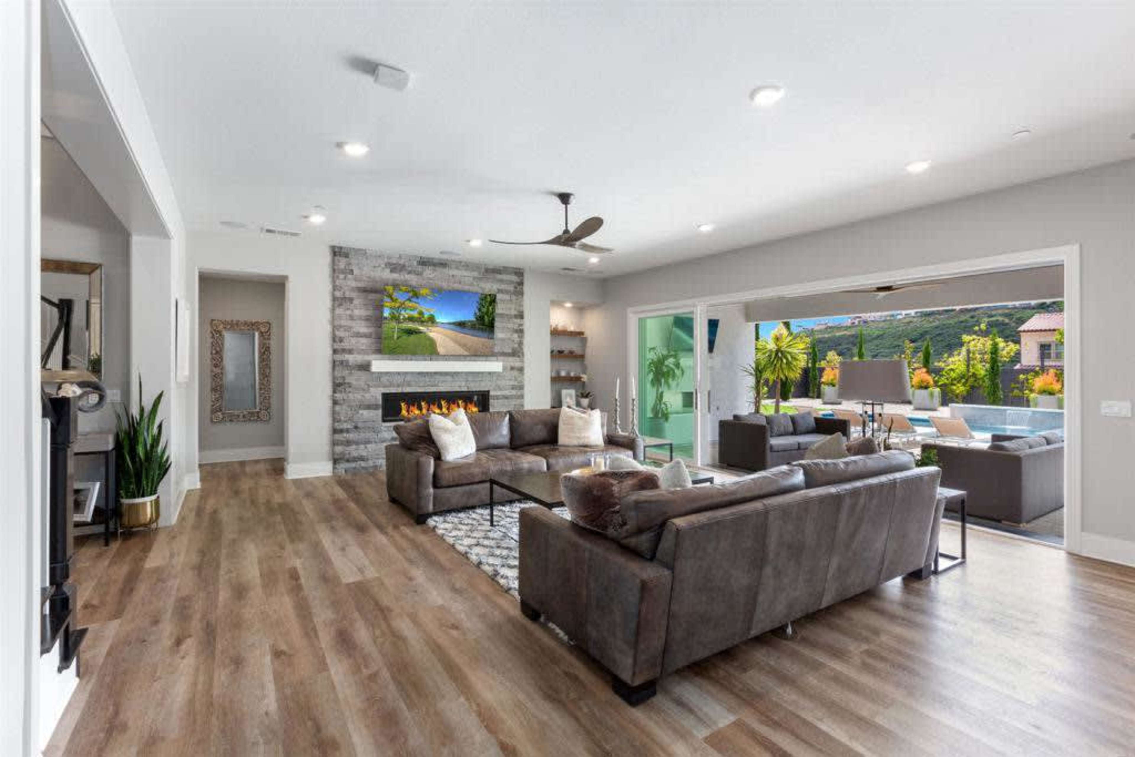 A modern living room features a stone fireplace, large windows showcasing an outdoor area, and sleek furniture arranged around a central rug.