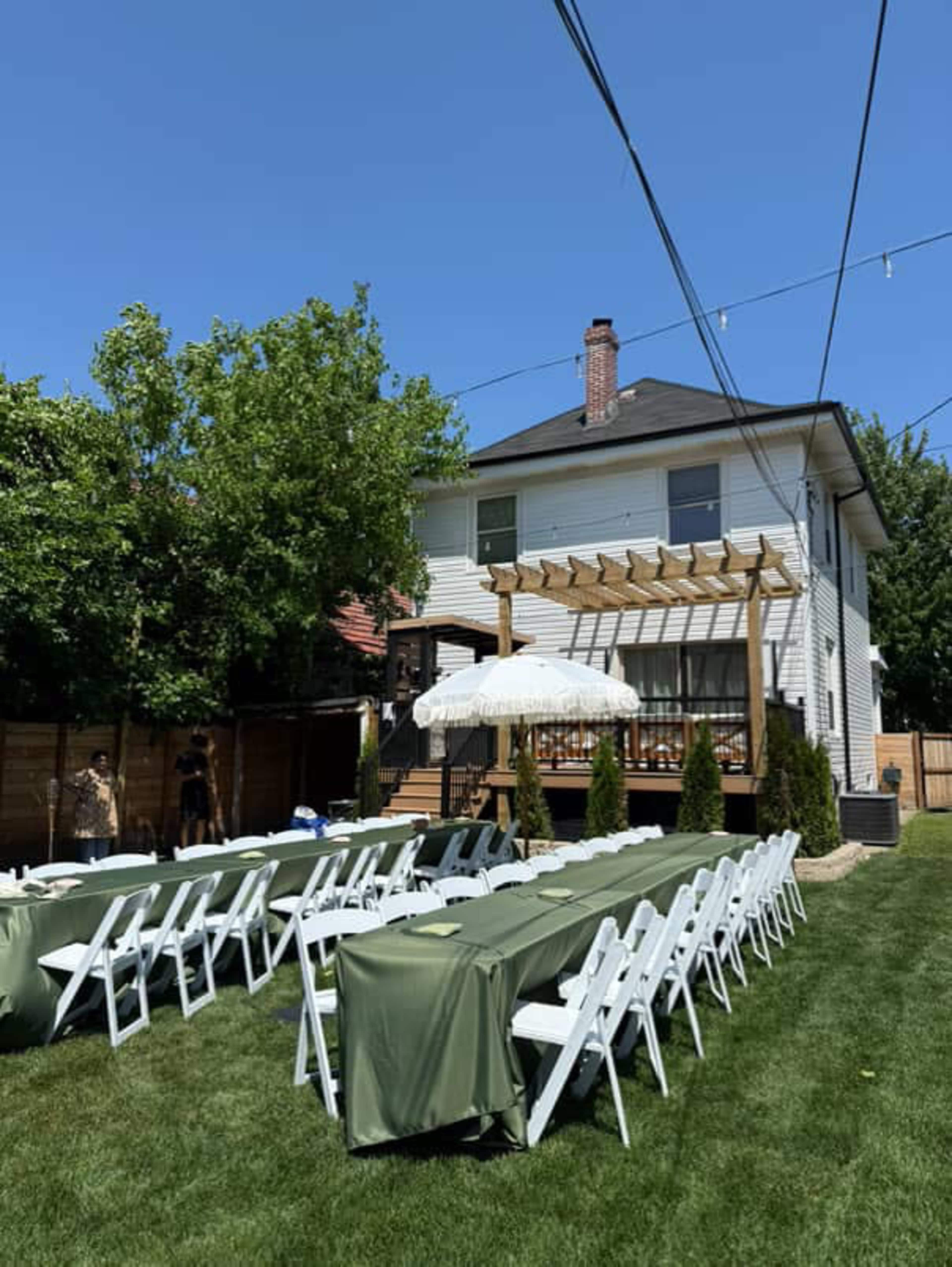 A backyard setup features rows of white chairs and tables covered with green tablecloths in front of a two-story house with a deck and an umbrella.