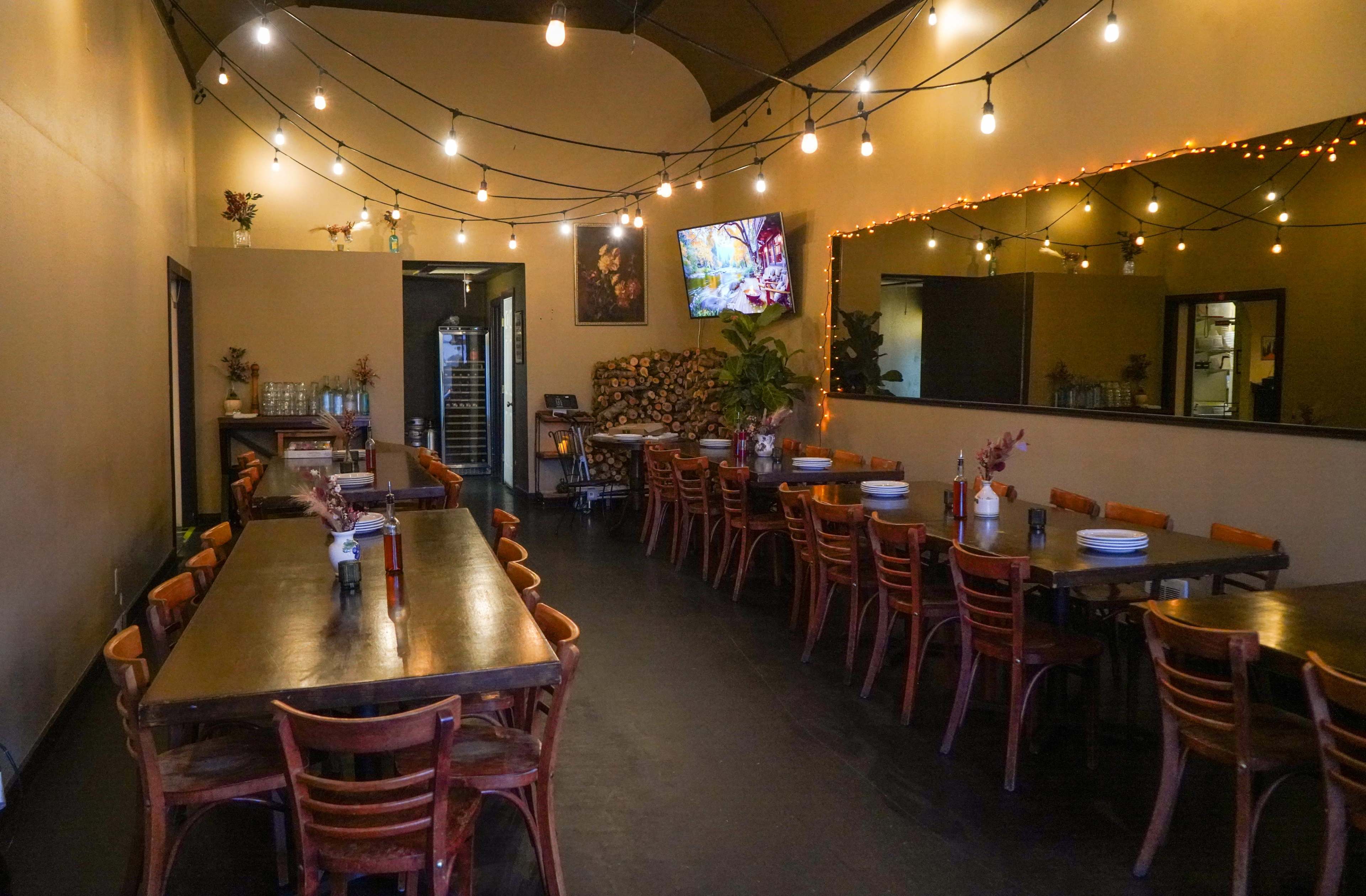 The image shows a restaurant interior with wooden tables and chairs, string lights hanging from the ceiling, and a television mounted on the wall.