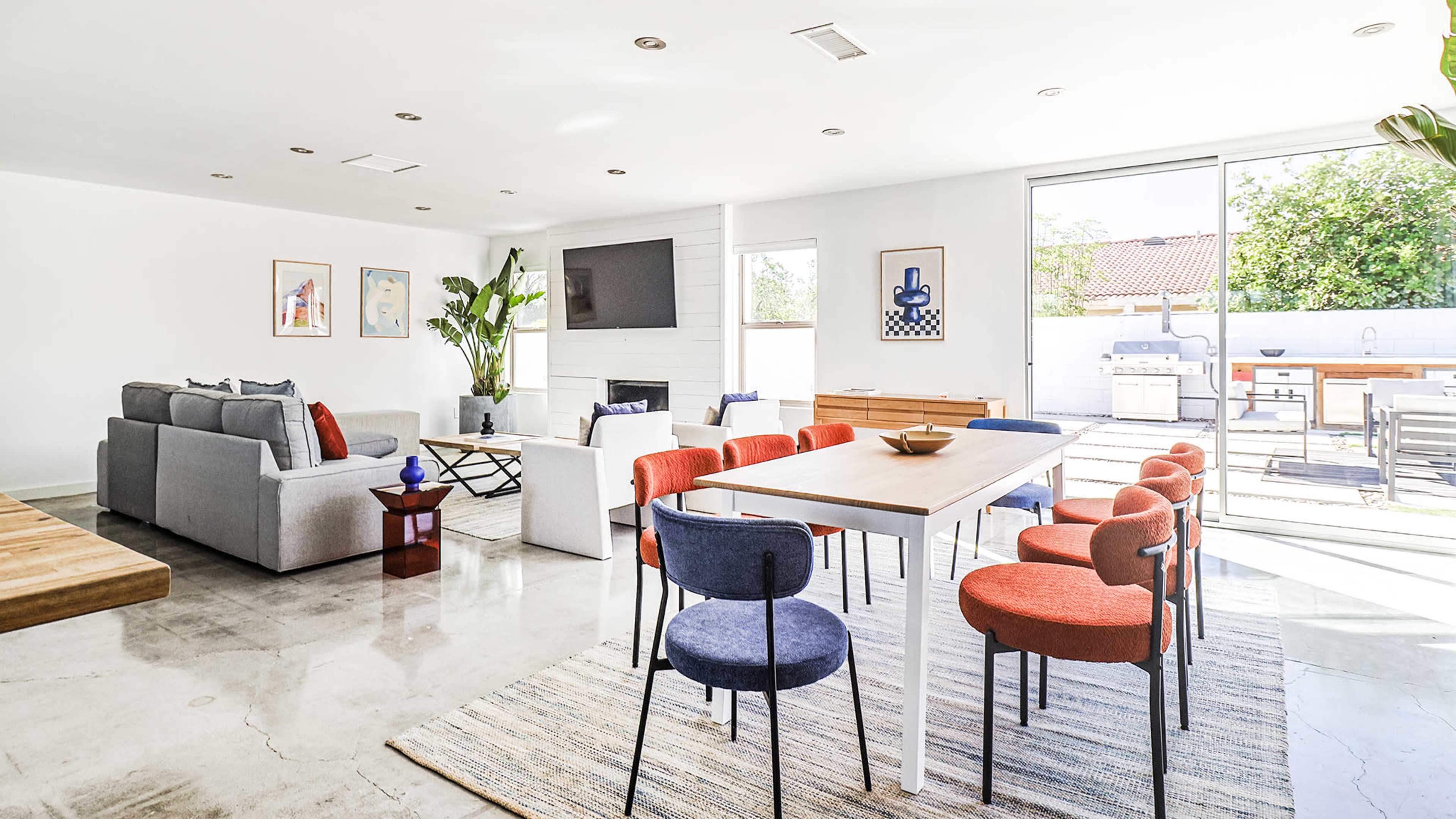 A modern, open-concept living and dining area featuring a sofa, a dining table with colorful chairs, and large windows leading to an outdoor space.