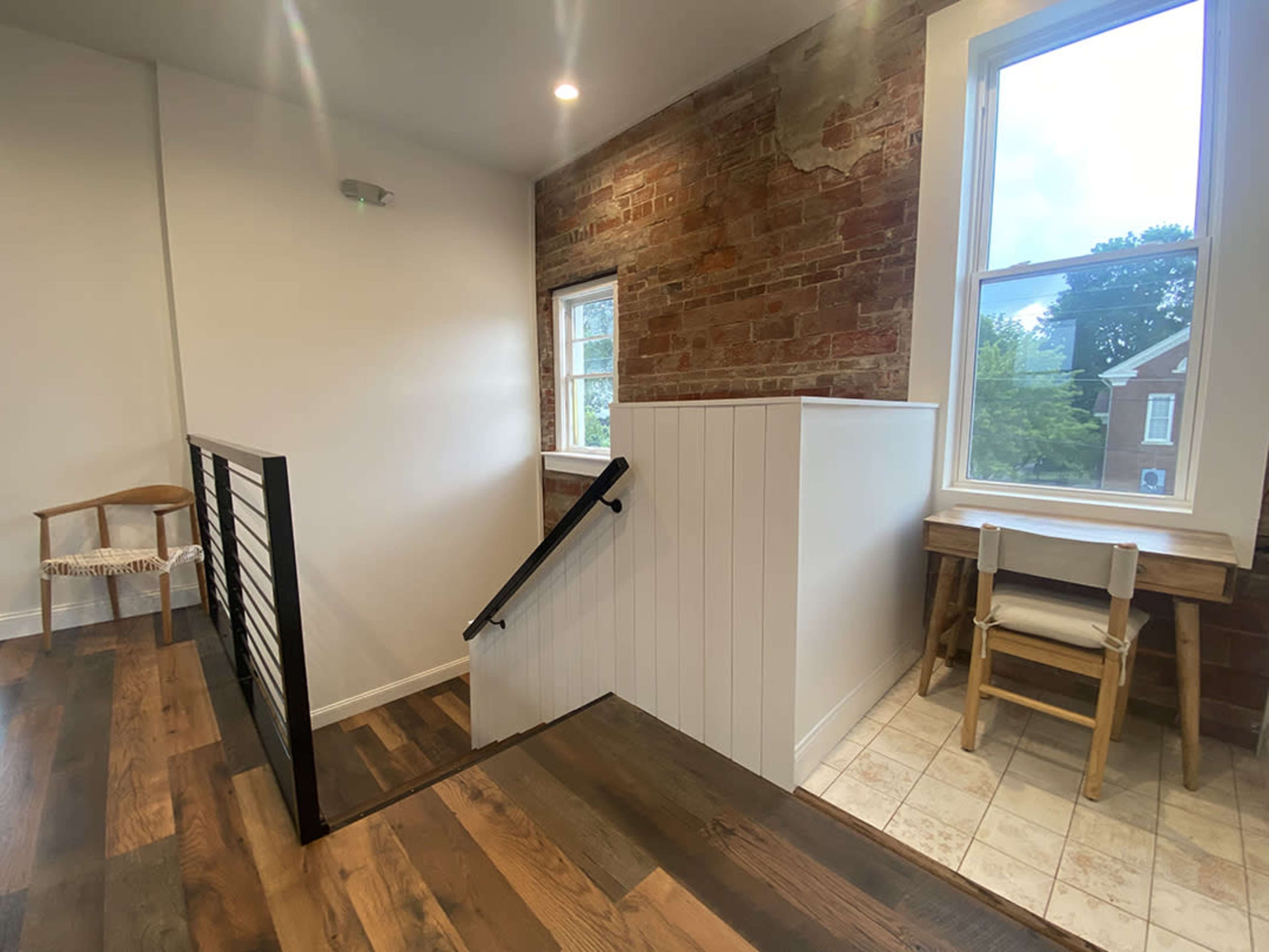 A well-lit interior space featuring a combination of exposed brick and white walls, with wooden flooring and a staircase leading to an upper level.