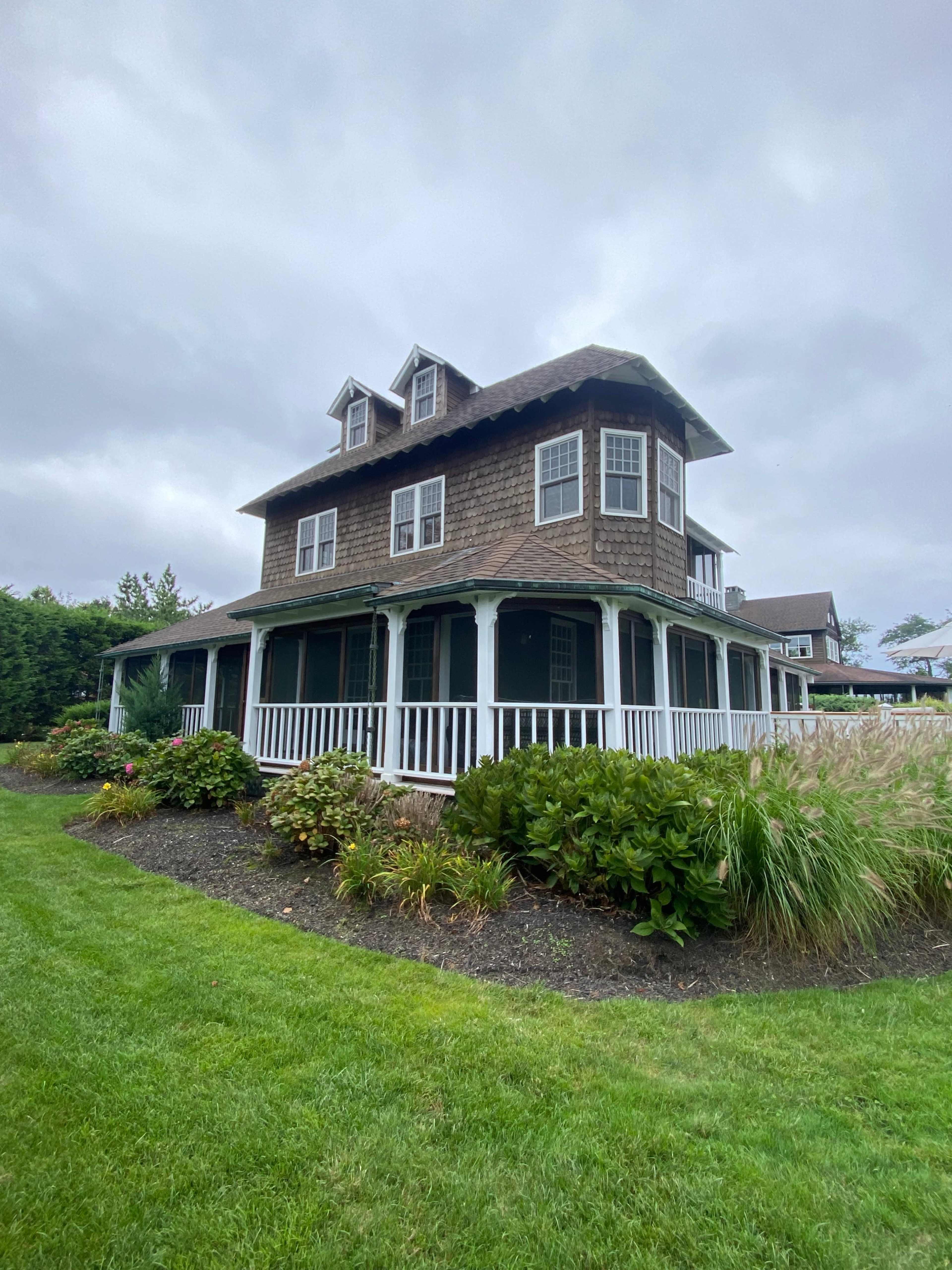 A three-story house with a shingled exterior and a wrap-around porch is surrounded by greenery on a cloudy day.