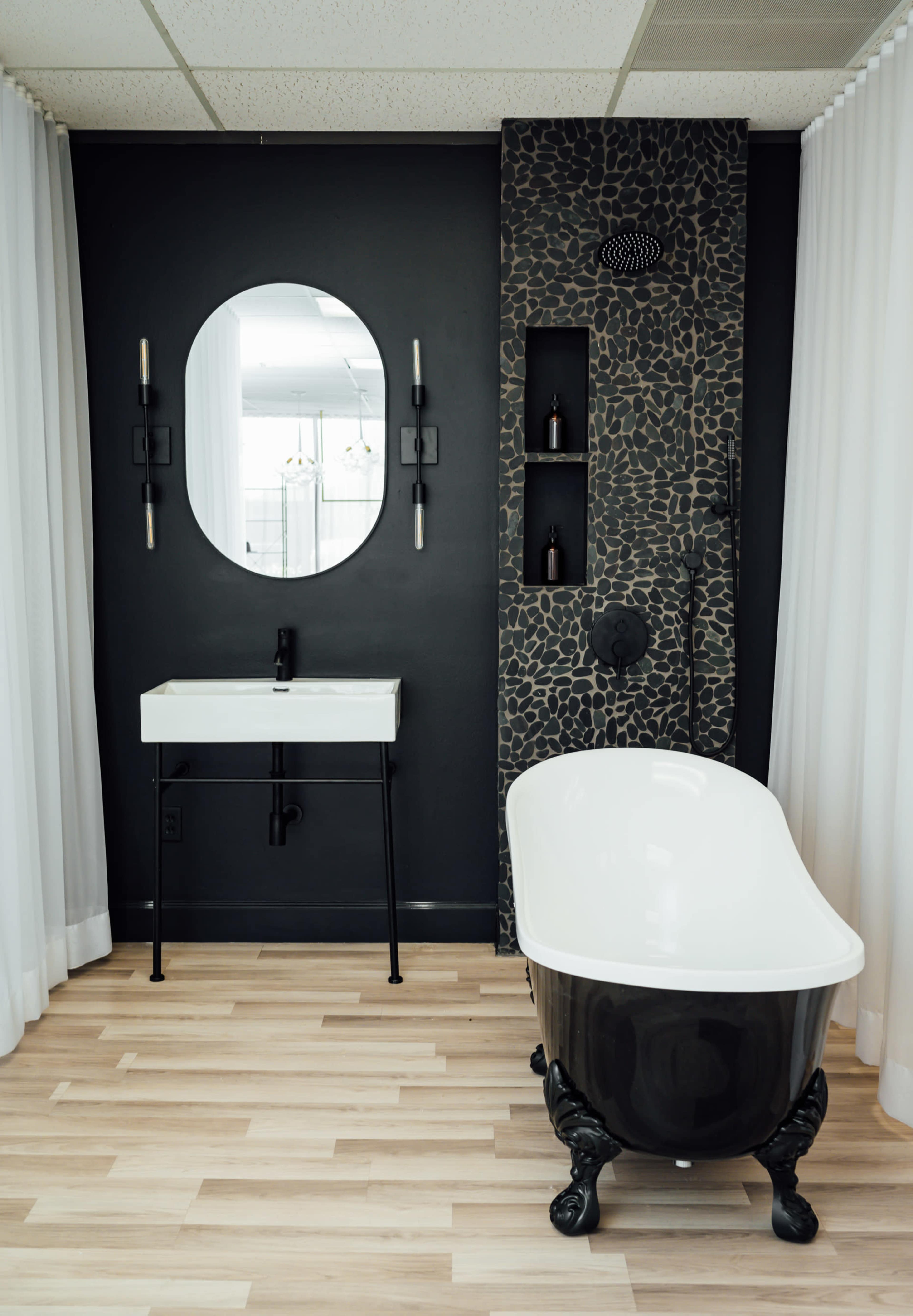 A modern bathroom featuring a freestanding black and white clawfoot bathtub, a minimalist sink with a sleek black faucet, and a dark wall with shelves for toiletries.
