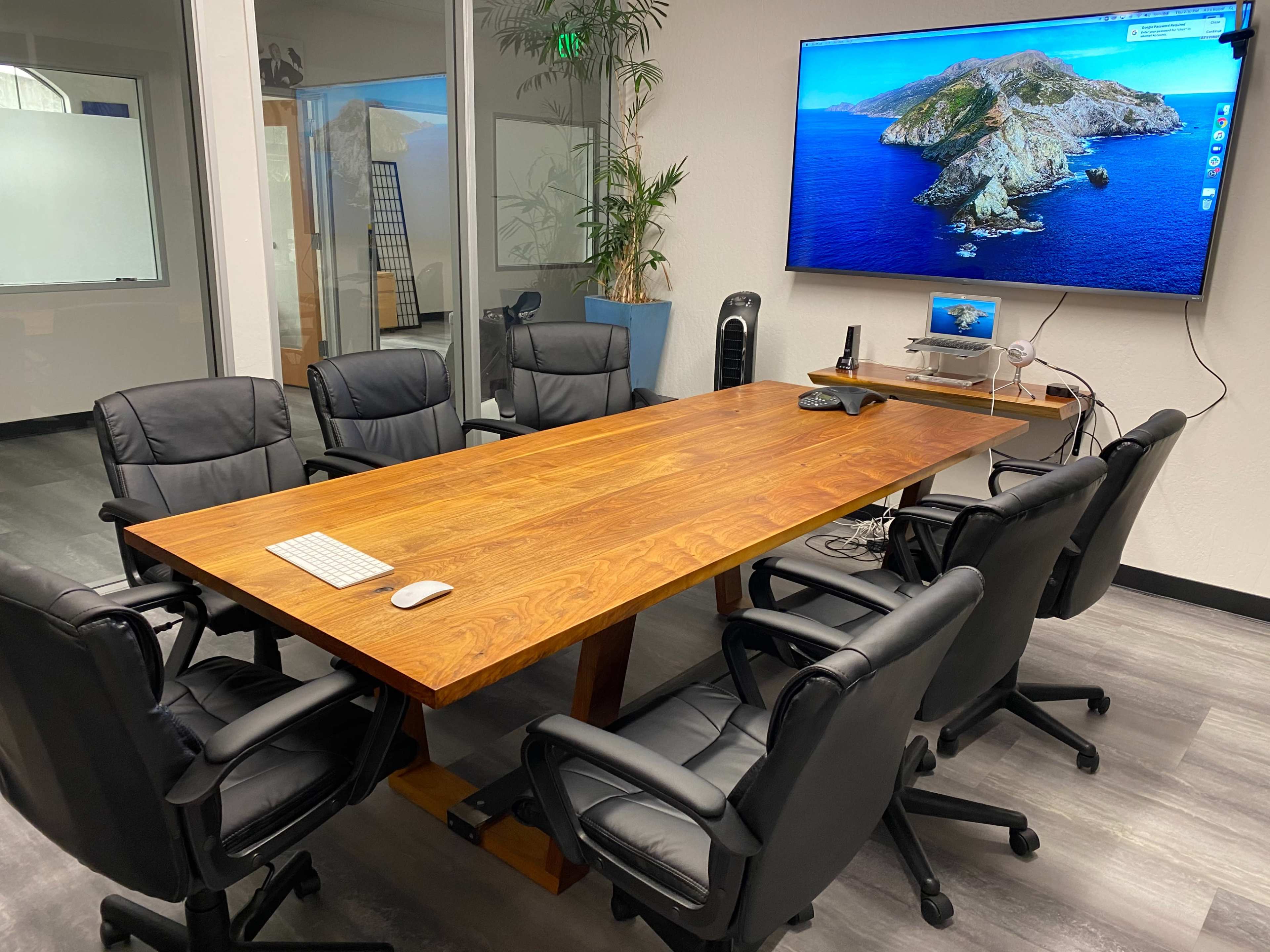 A meeting room features a large wooden table surrounded by six black office chairs, with a wall-mounted screen displaying a coastal image.