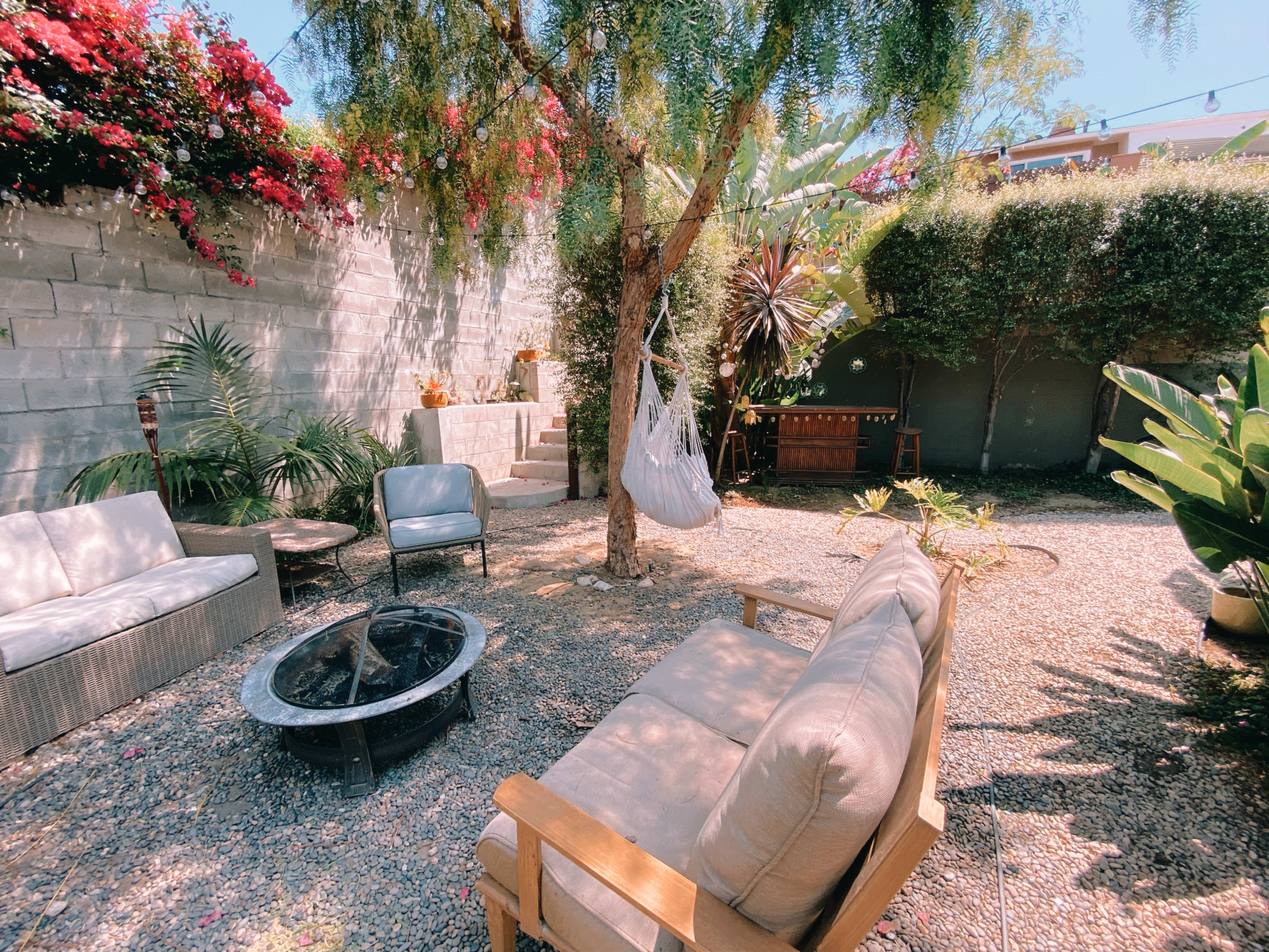 A cozy outdoor garden space features seating around a fire pit, a hammock hanging from a tree, and lush greenery with colorful flowers.