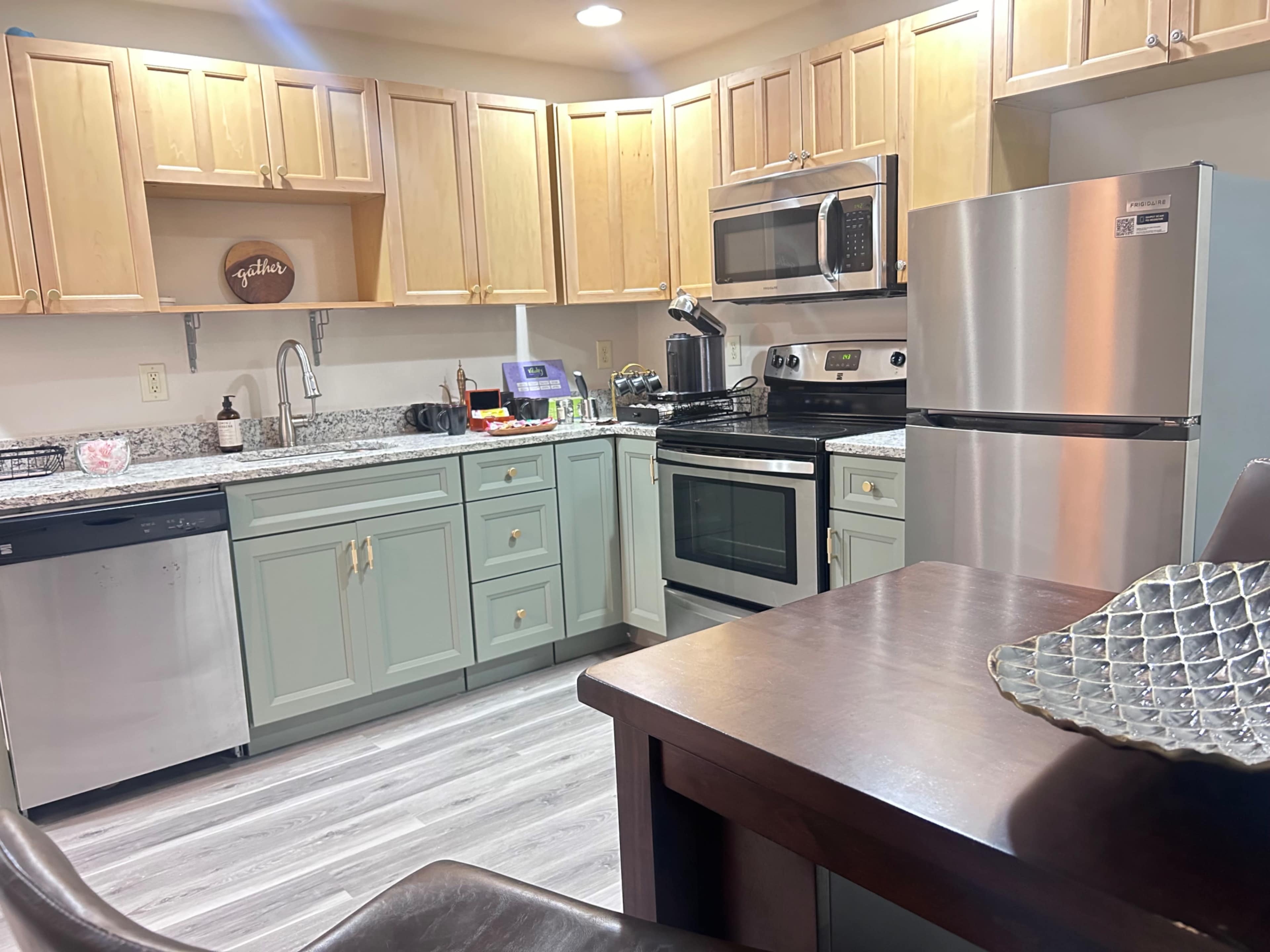 A modern kitchen features light wood cabinetry, stainless steel appliances, and gray countertops, with a dining table in the foreground.