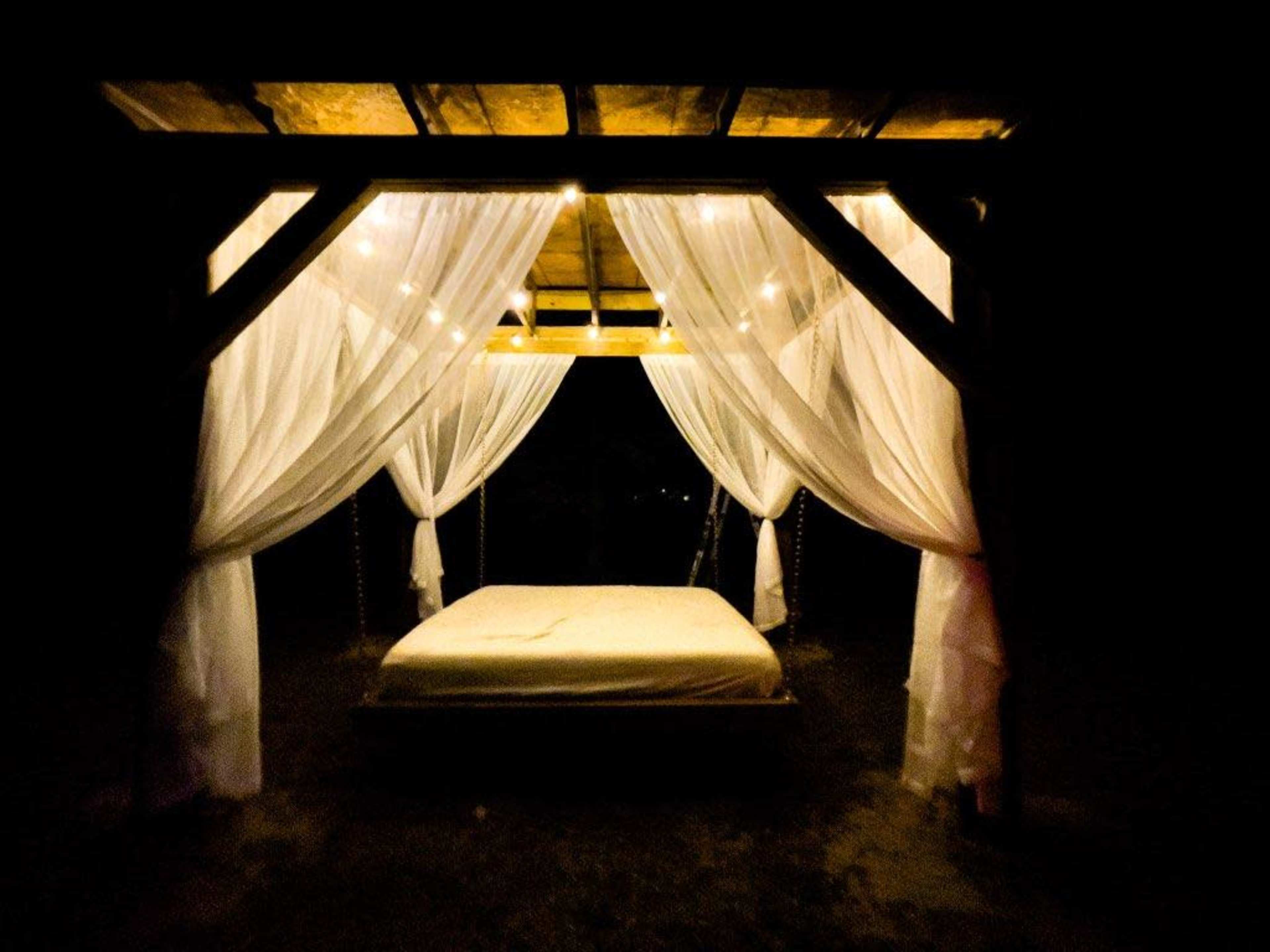 A framed bed with sheer white drapes is illuminated by soft lights in a dark setting.
