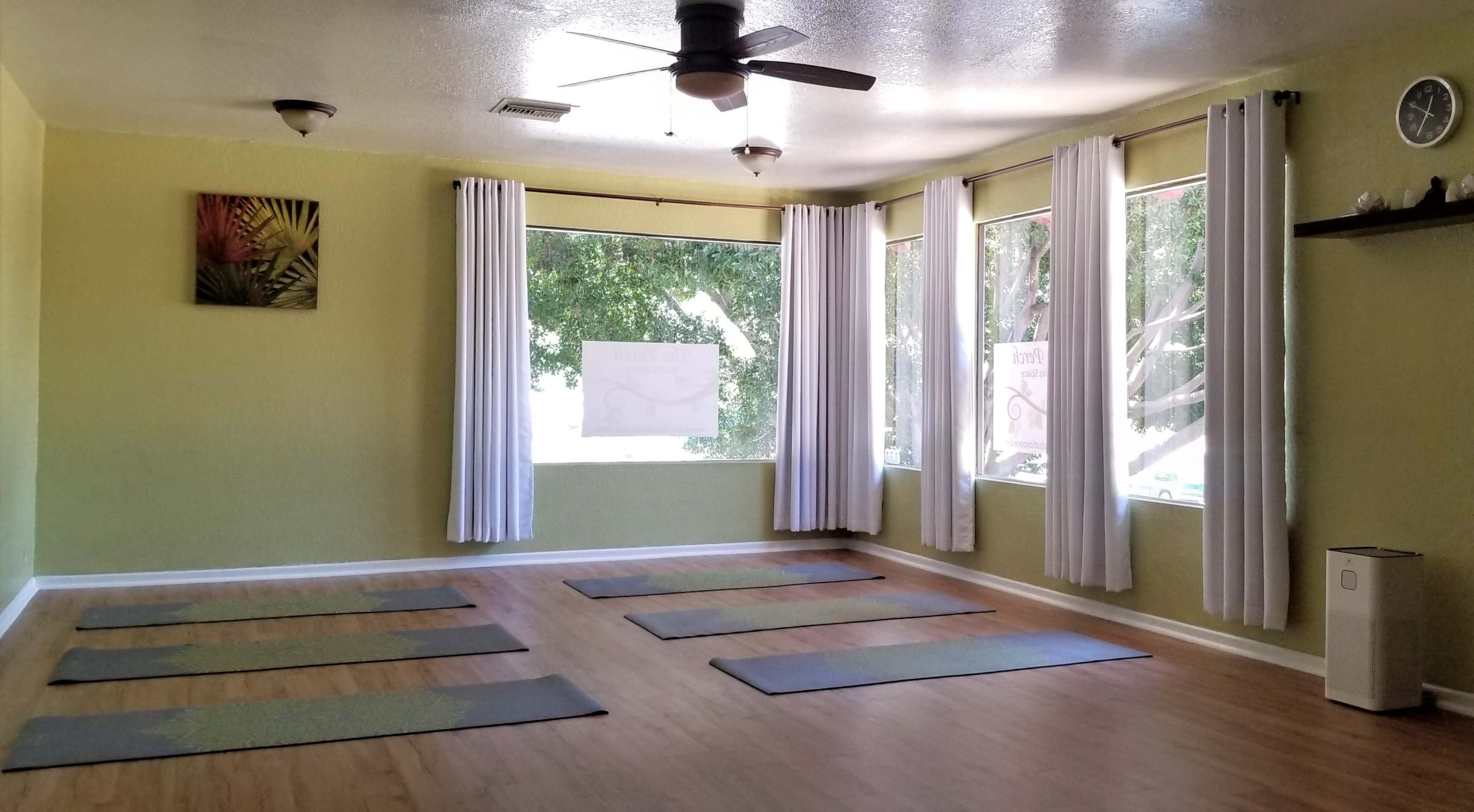 A spacious yoga studio with wooden flooring, several yoga mats arranged on the floor, and large windows letting in natural light.
