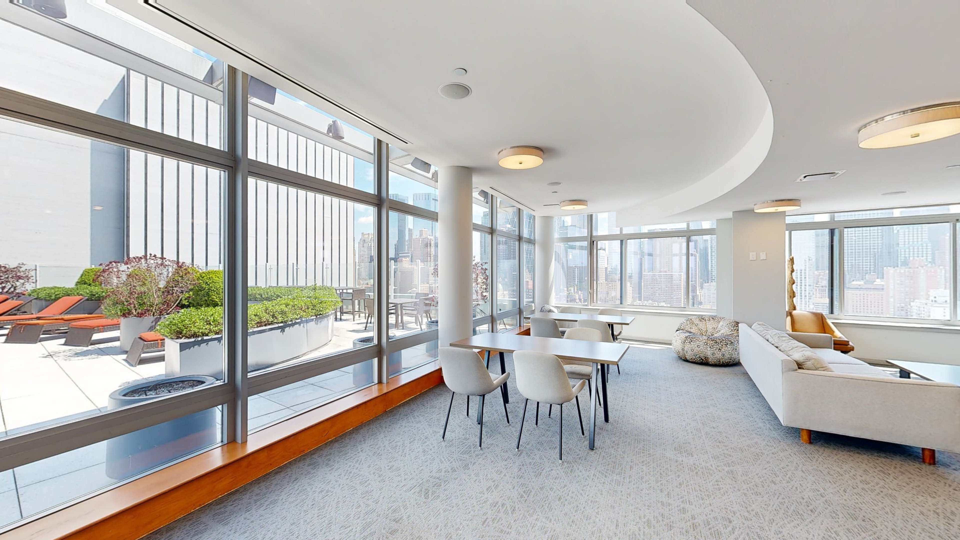 A modern lounge space with large windows, featuring seating arrangements and views of an outdoor terrace in a high-rise building.