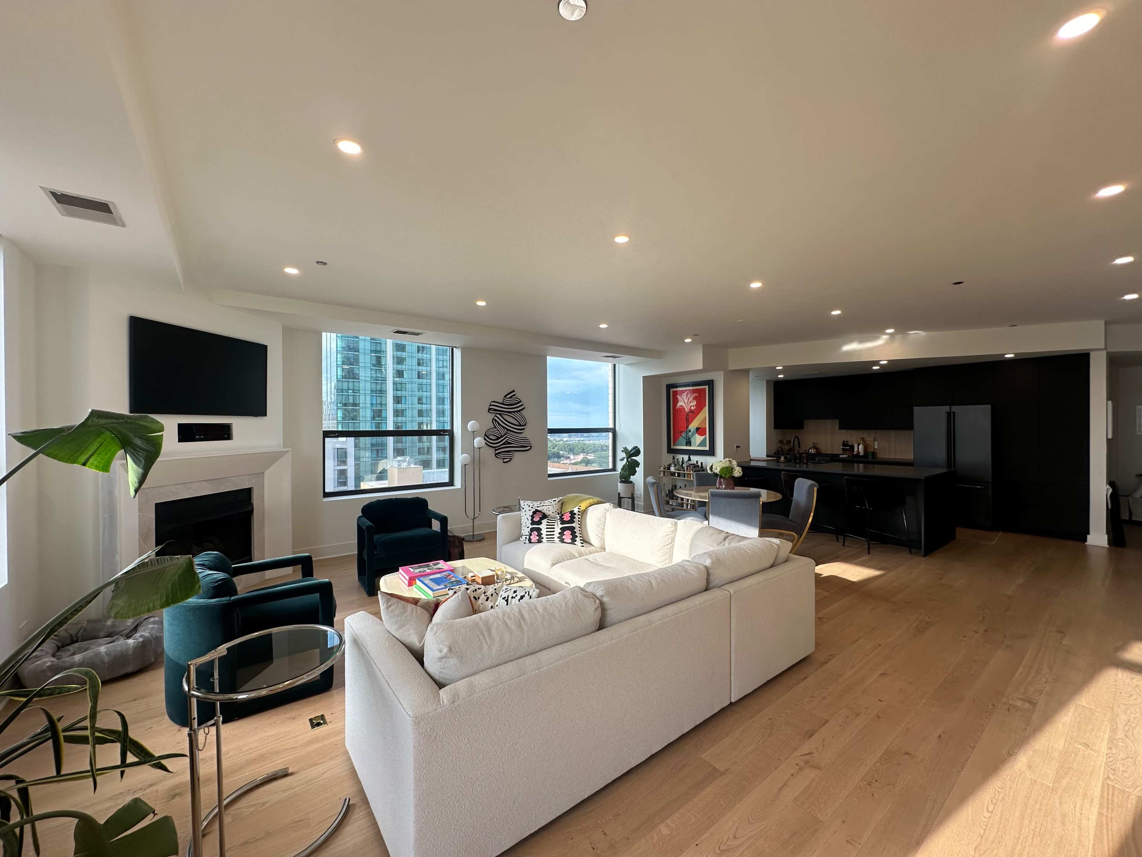 A modern living area with a light-colored sofa, two teal chairs, a coffee table, and an open kitchen featuring black cabinetry and a large window with a view.