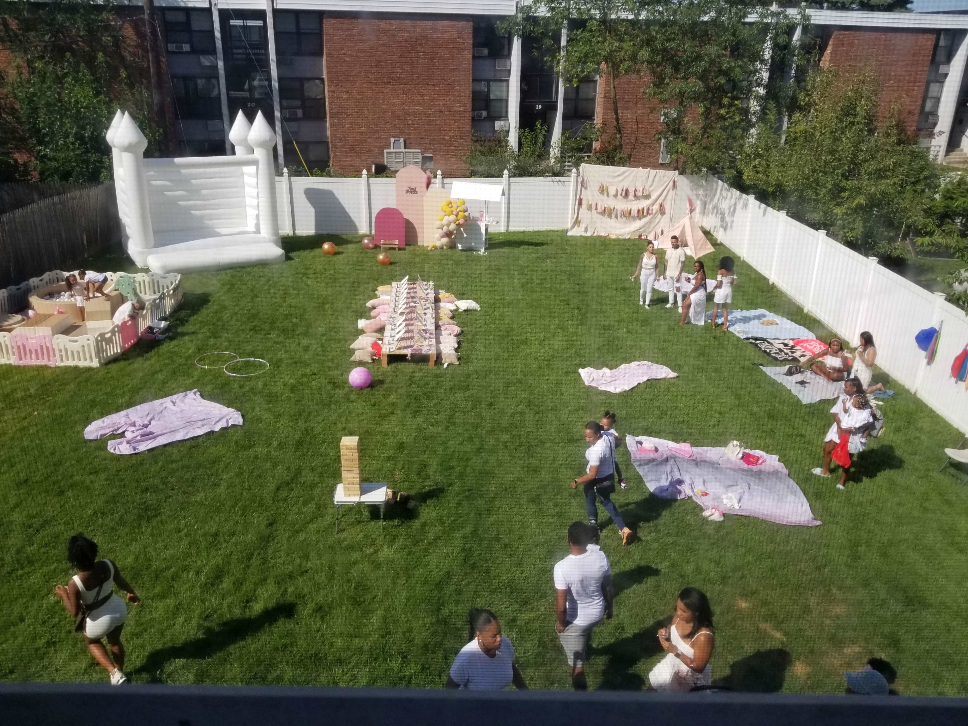 The image shows a large grassy backyard setup for a party, featuring a bounce house, picnic seating, and various decorations while people are gathered around.