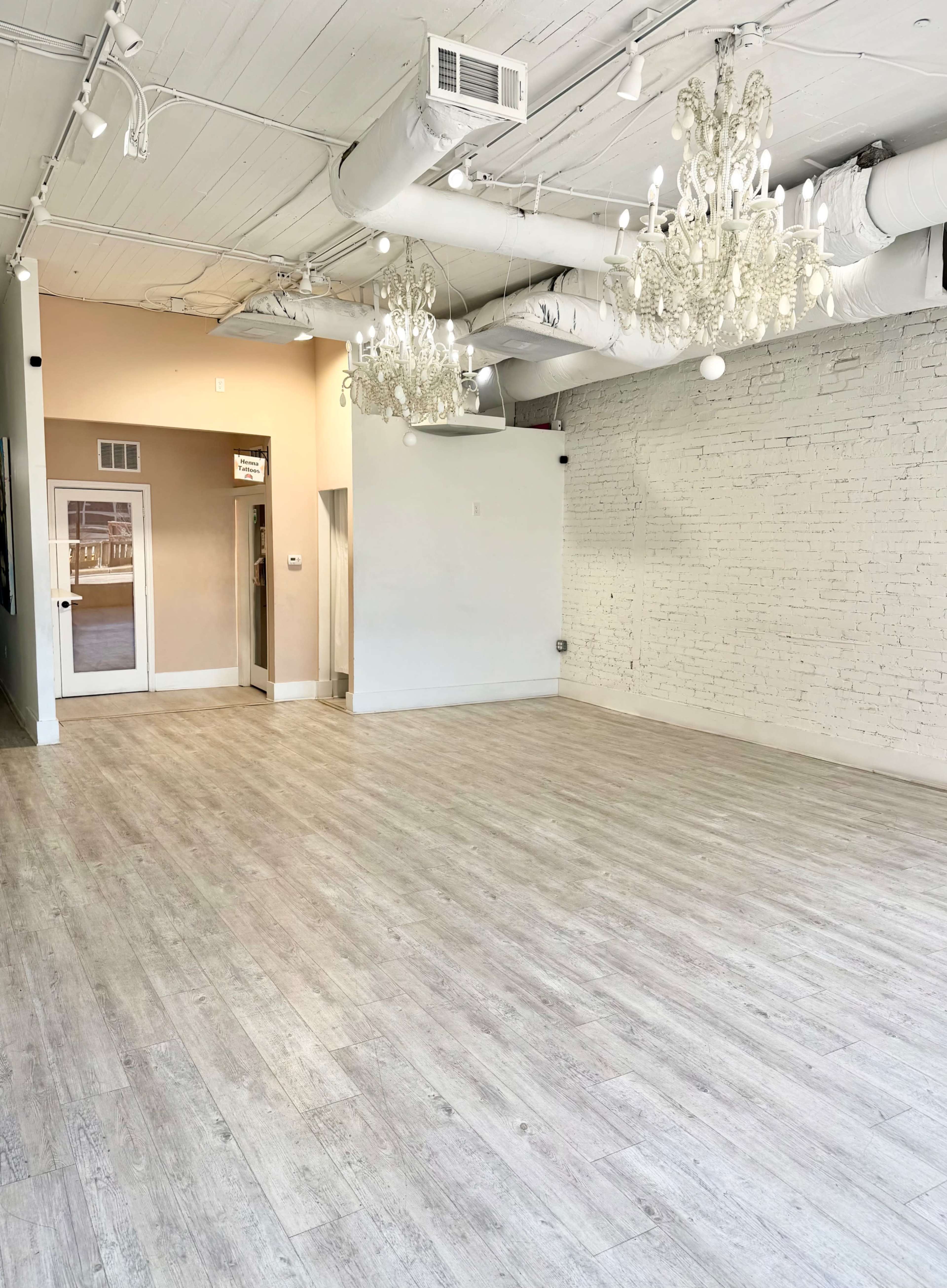 The image shows a spacious, bright room with wooden flooring, white brick walls, and two chandeliers hanging from the ceiling.
