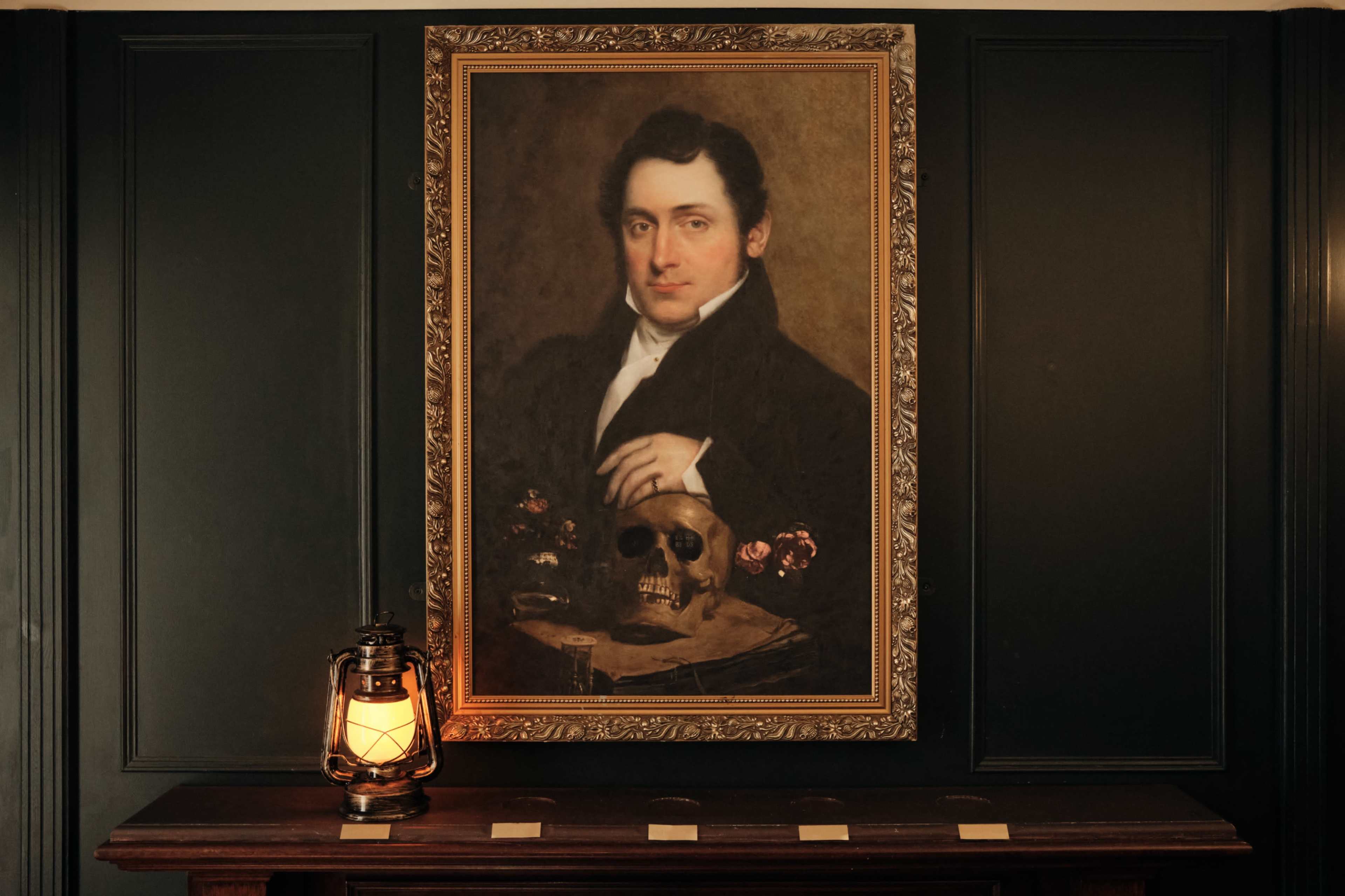 The image features a historic portrait of a man in a dark coat, framed and displayed above a wooden table with a skull and an oil lantern, against a backdrop of dark walls.