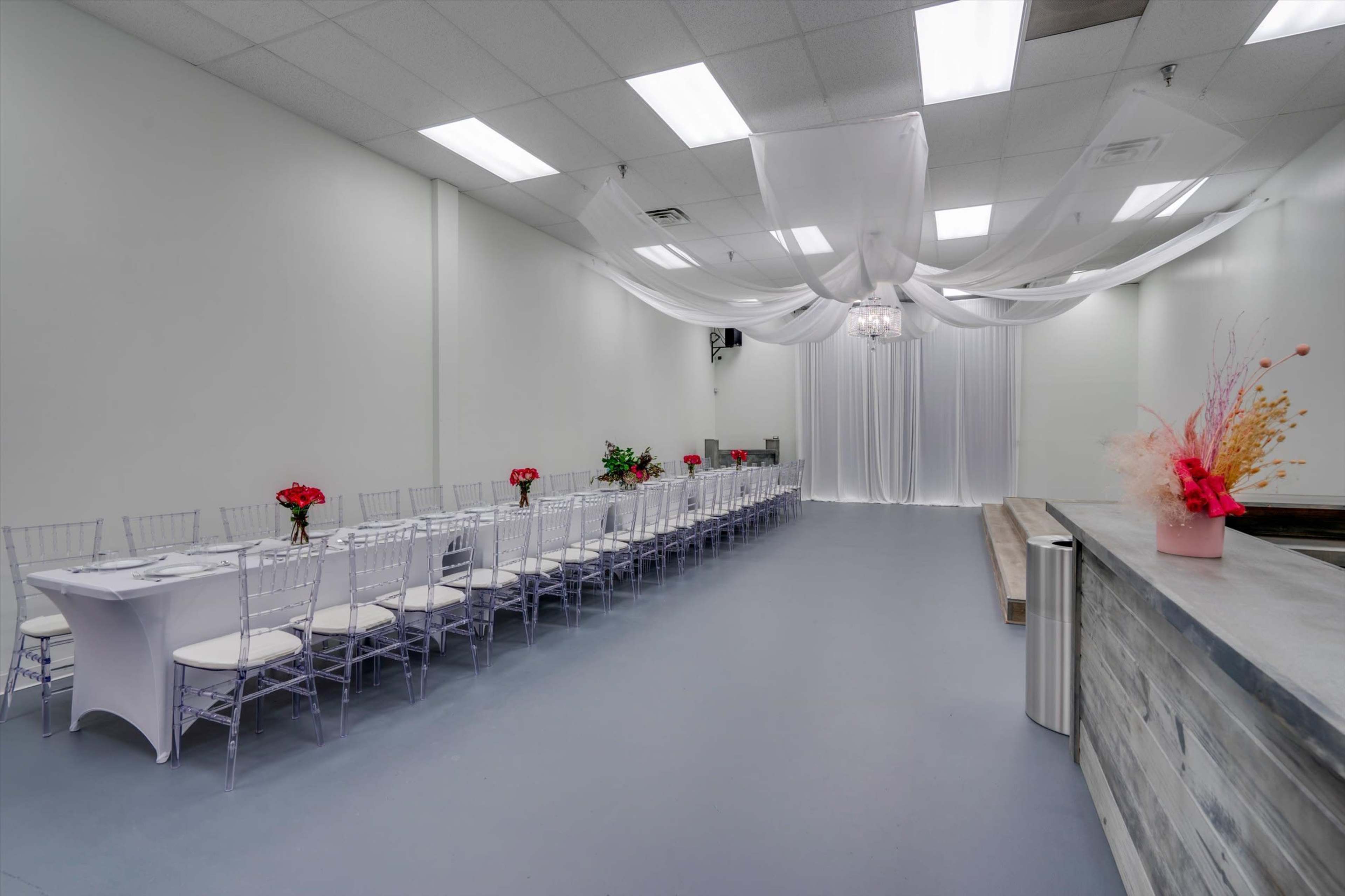 A spacious event room features a long table set for dining, adorned with floral centerpieces, and draped fabric hanging from the ceiling.