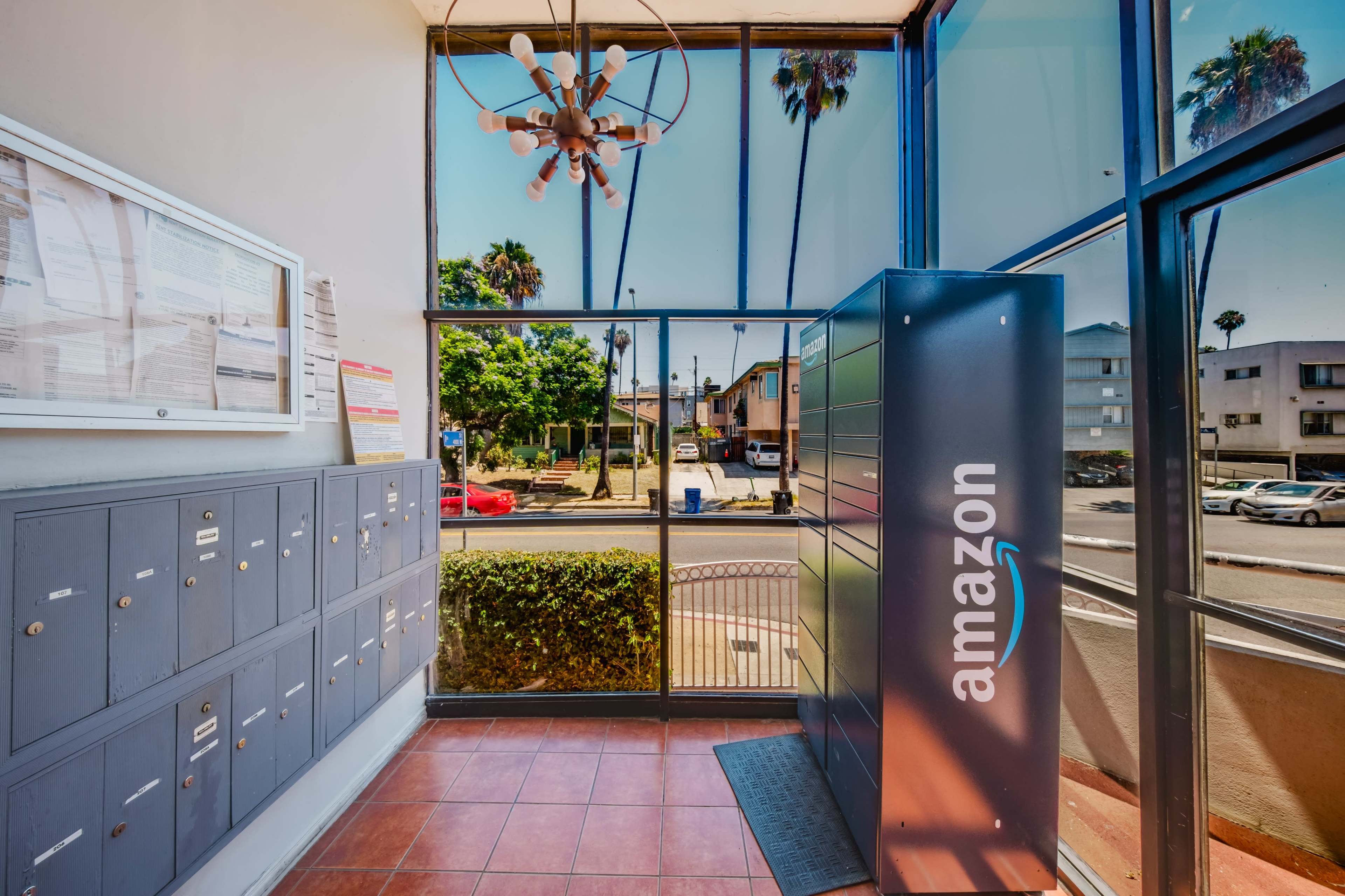 The image shows a lobby area with a row of mailboxes on the left and an Amazon package locker on the right, with windows providing a view of palm trees and an exterior street.