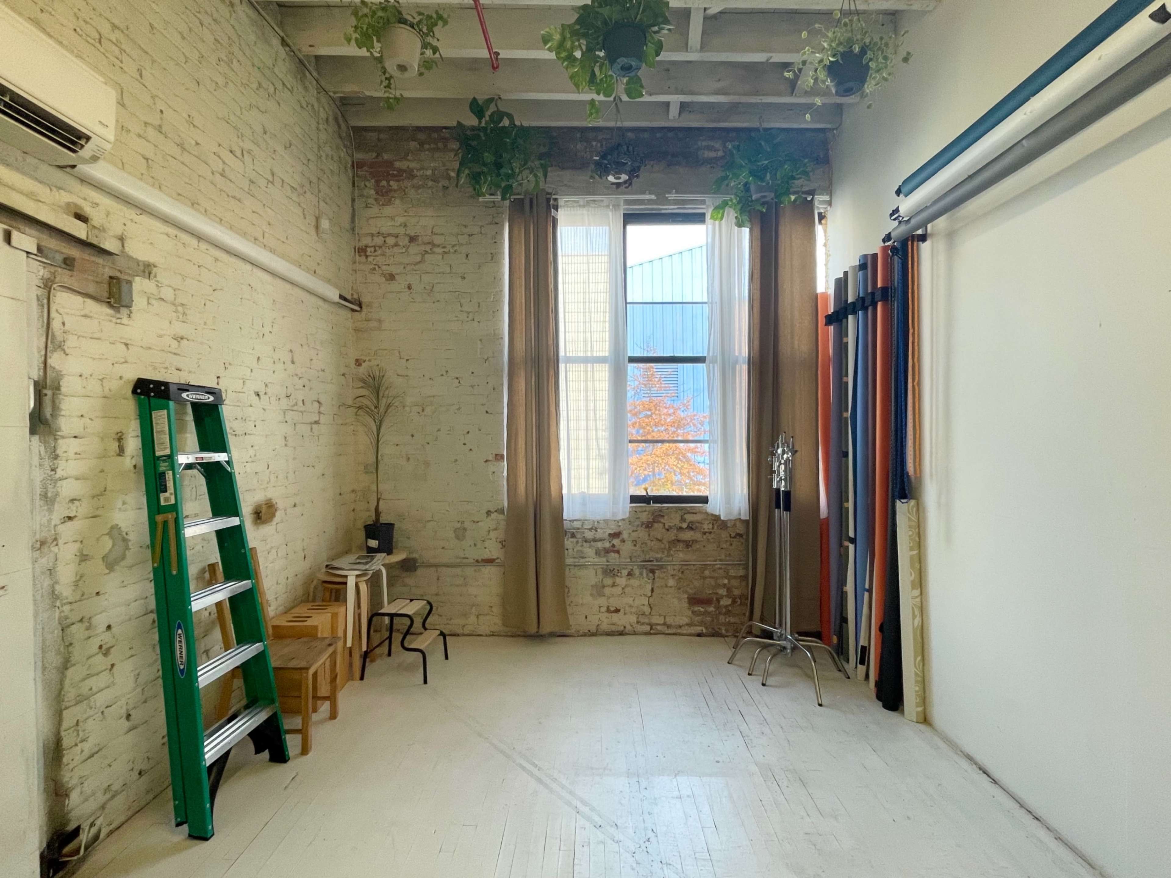 The image shows a spacious, empty room with exposed brick walls, a large window, and a ladder, along with a few chairs and draped fabrics.
