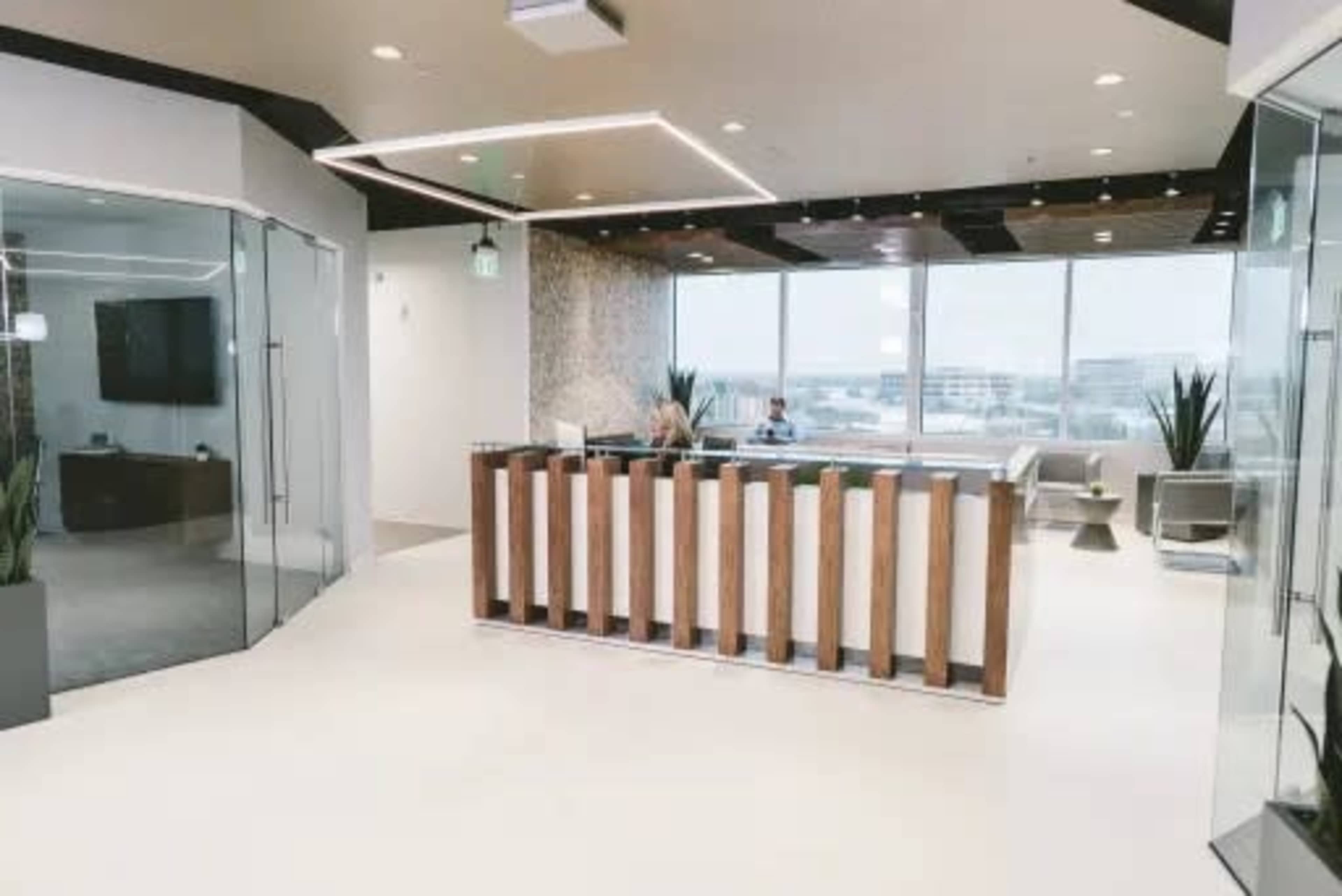 A modern office reception area with a wood-paneled front desk, large windows, and minimalist furniture.