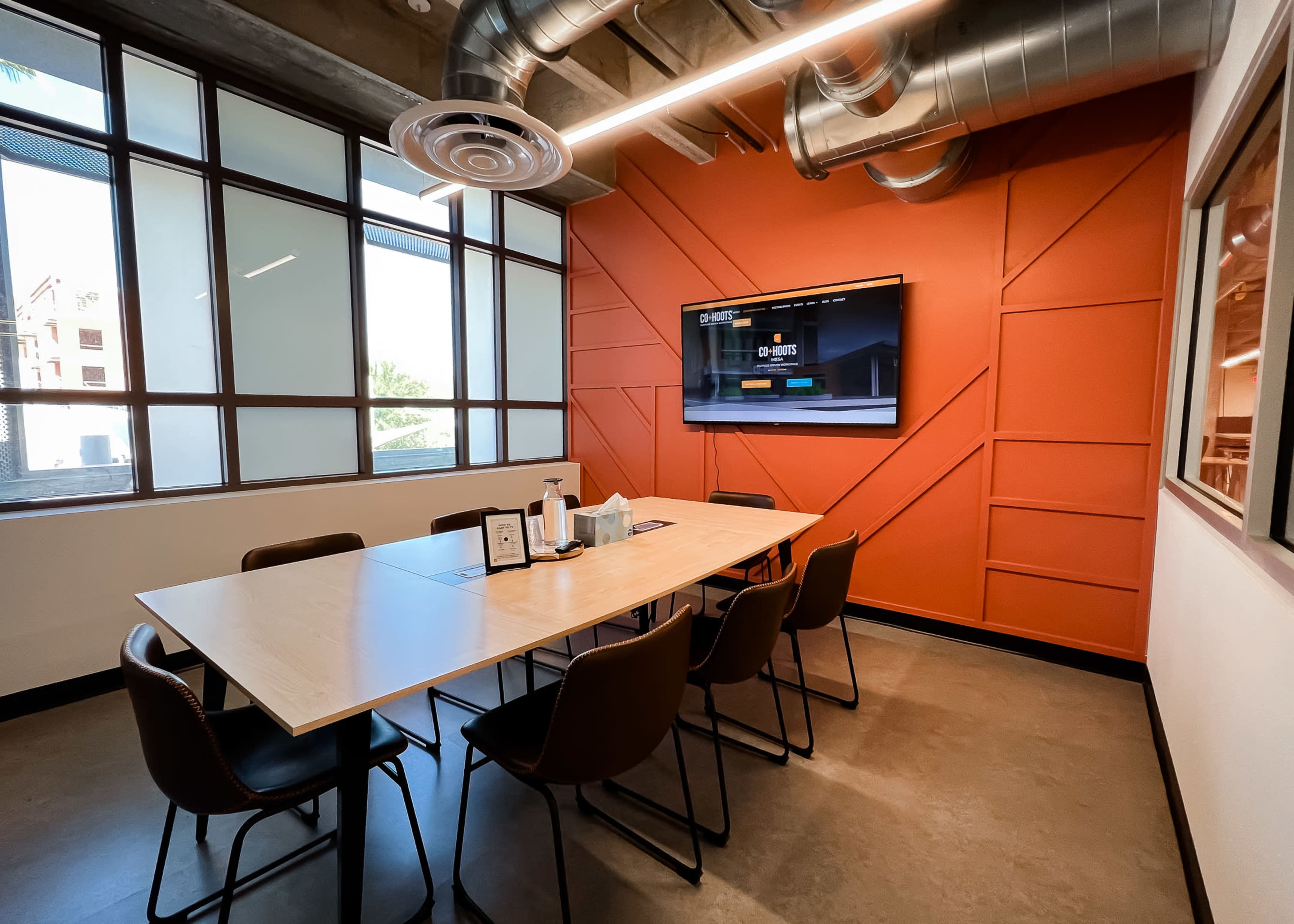 A conference room features a large table surrounded by chairs, with a television mounted on an orange accent wall and large windows providing natural light.