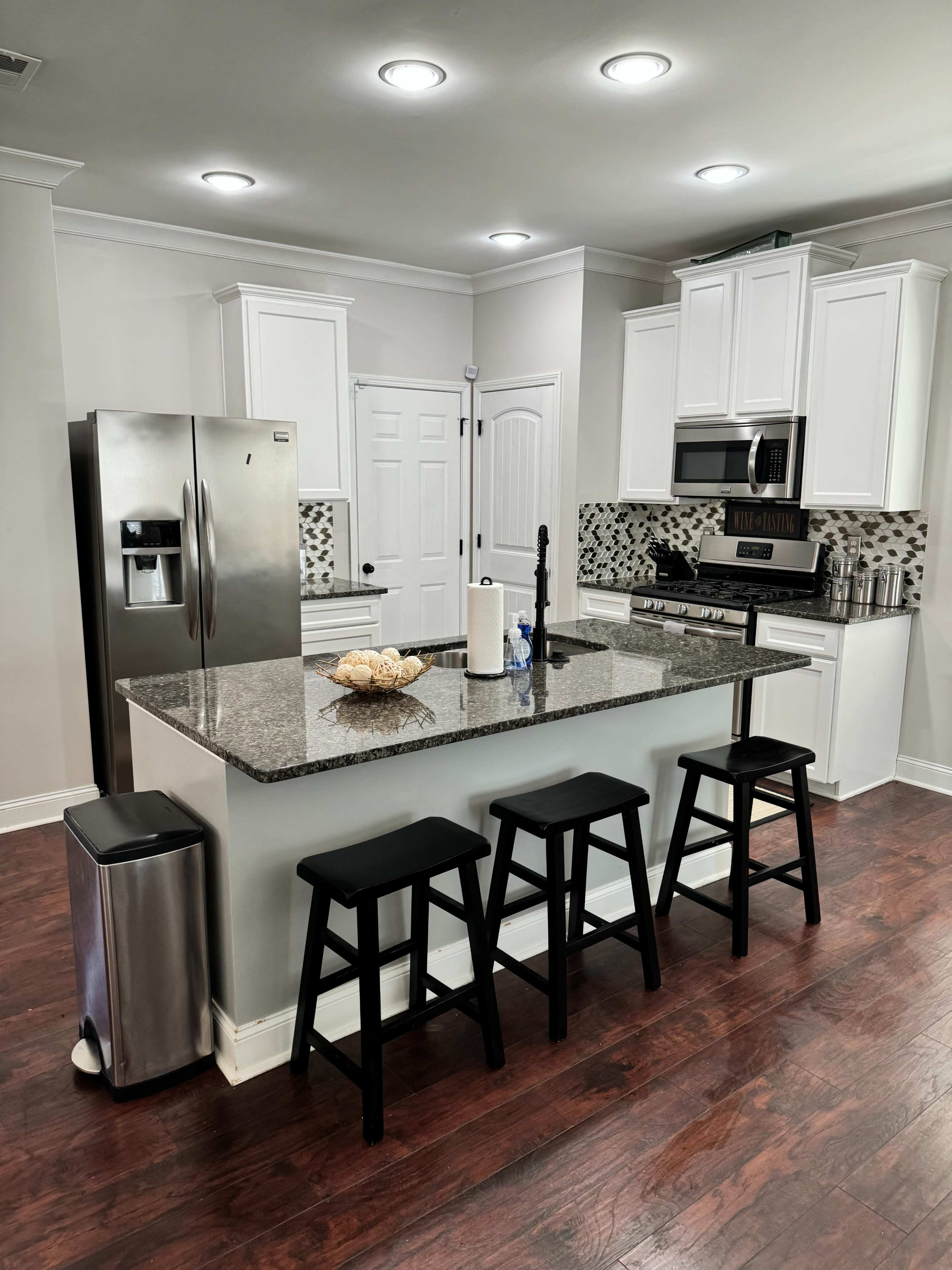 The image shows a modern kitchen with white cabinets, a gray granite island with three black stools, stainless steel appliances, and wooden flooring.