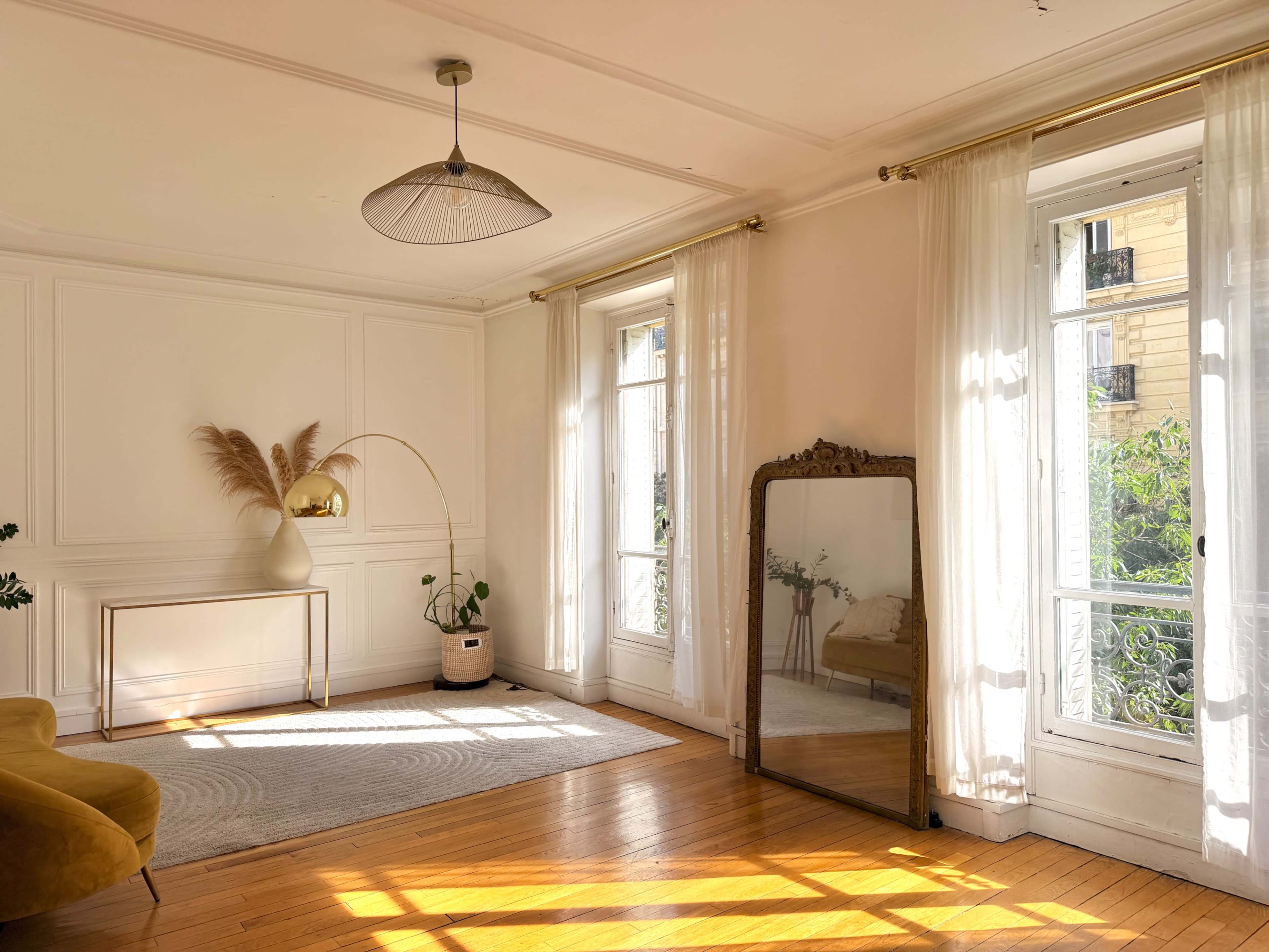 The bright room features large windows with sheer curtains, a gold-accented mirror, a decorative floor lamp, and a cozy yellow couch.