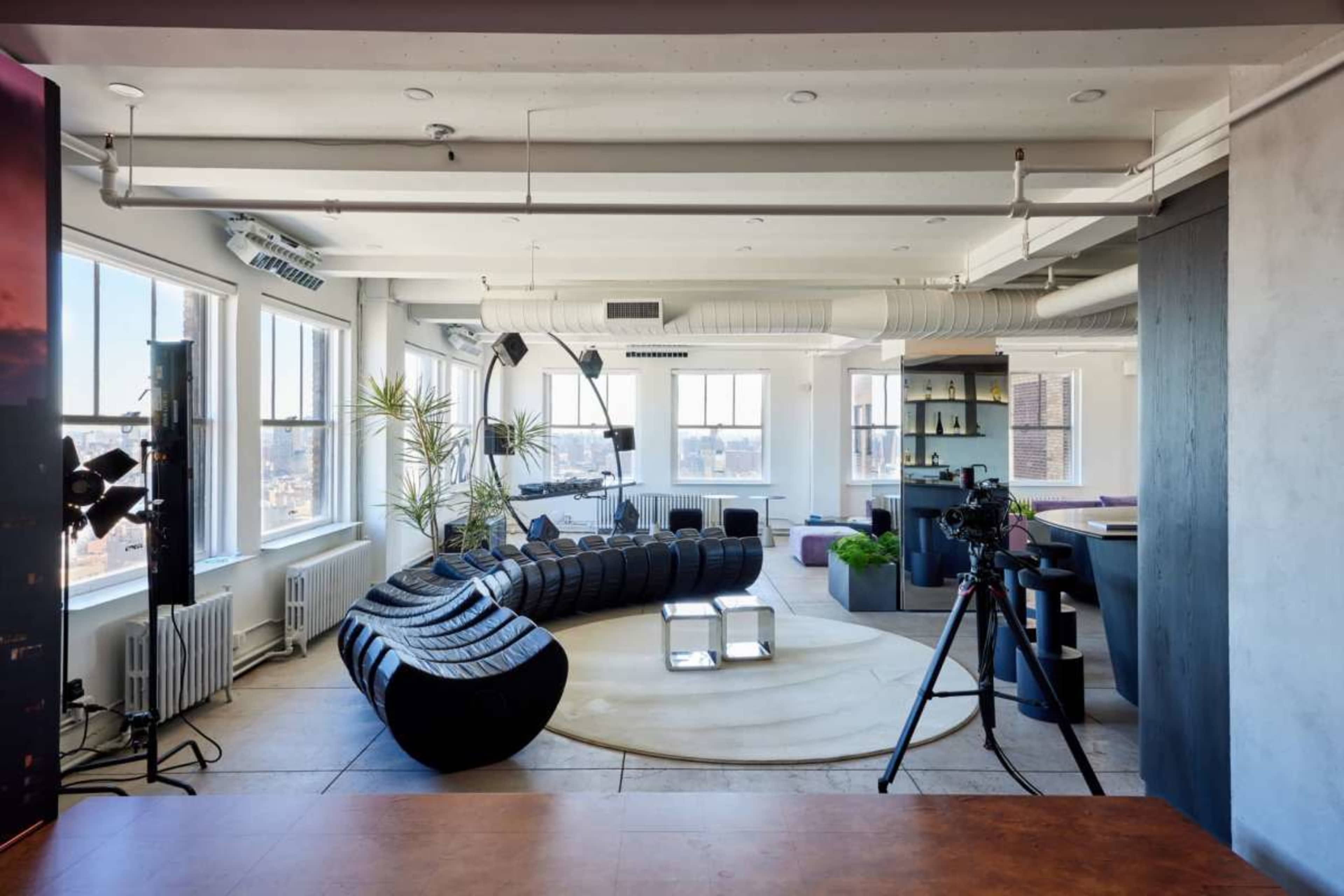 The image shows a modern interior of an open-plan studio with large windows, a circular area rug, a curved sofa, and various lighting equipment set up for a photoshoot.
