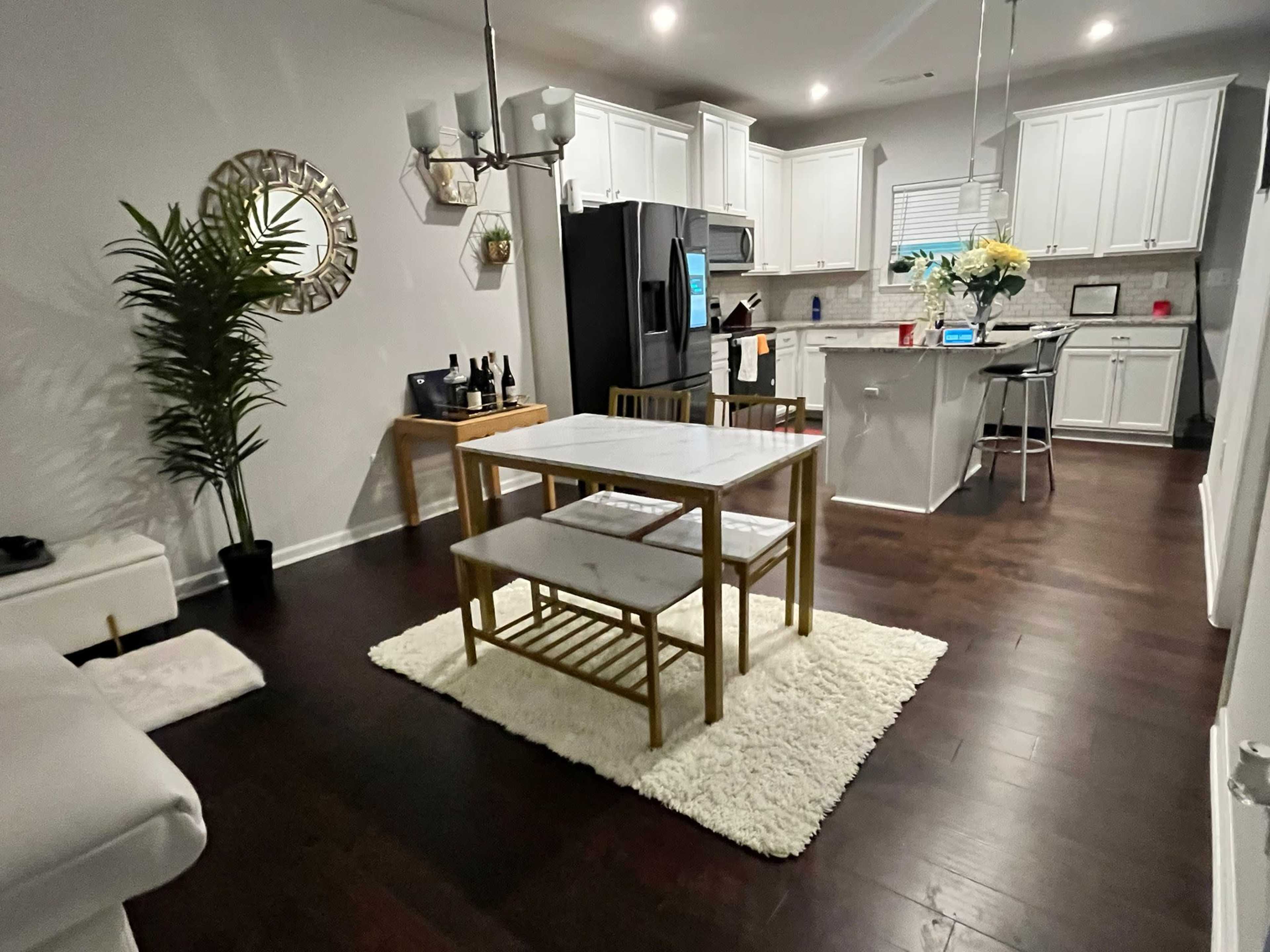 A modern kitchen and dining area features a white table with two benches on a rug, surrounded by cabinetry and appliances.