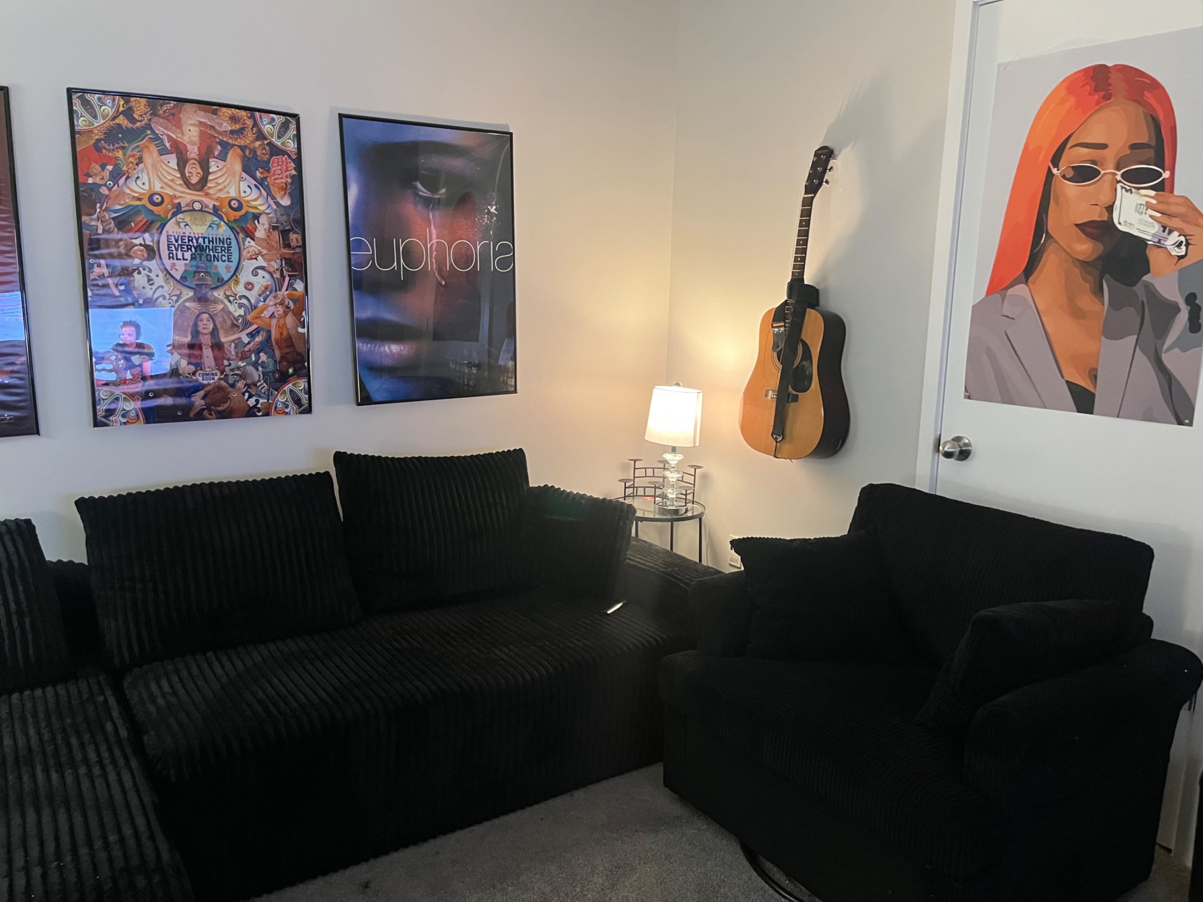 The scene features a cozy room with a black upholstered couch, a chair, several colorful posters on the walls, a guitar hanging, and a lamp providing light.