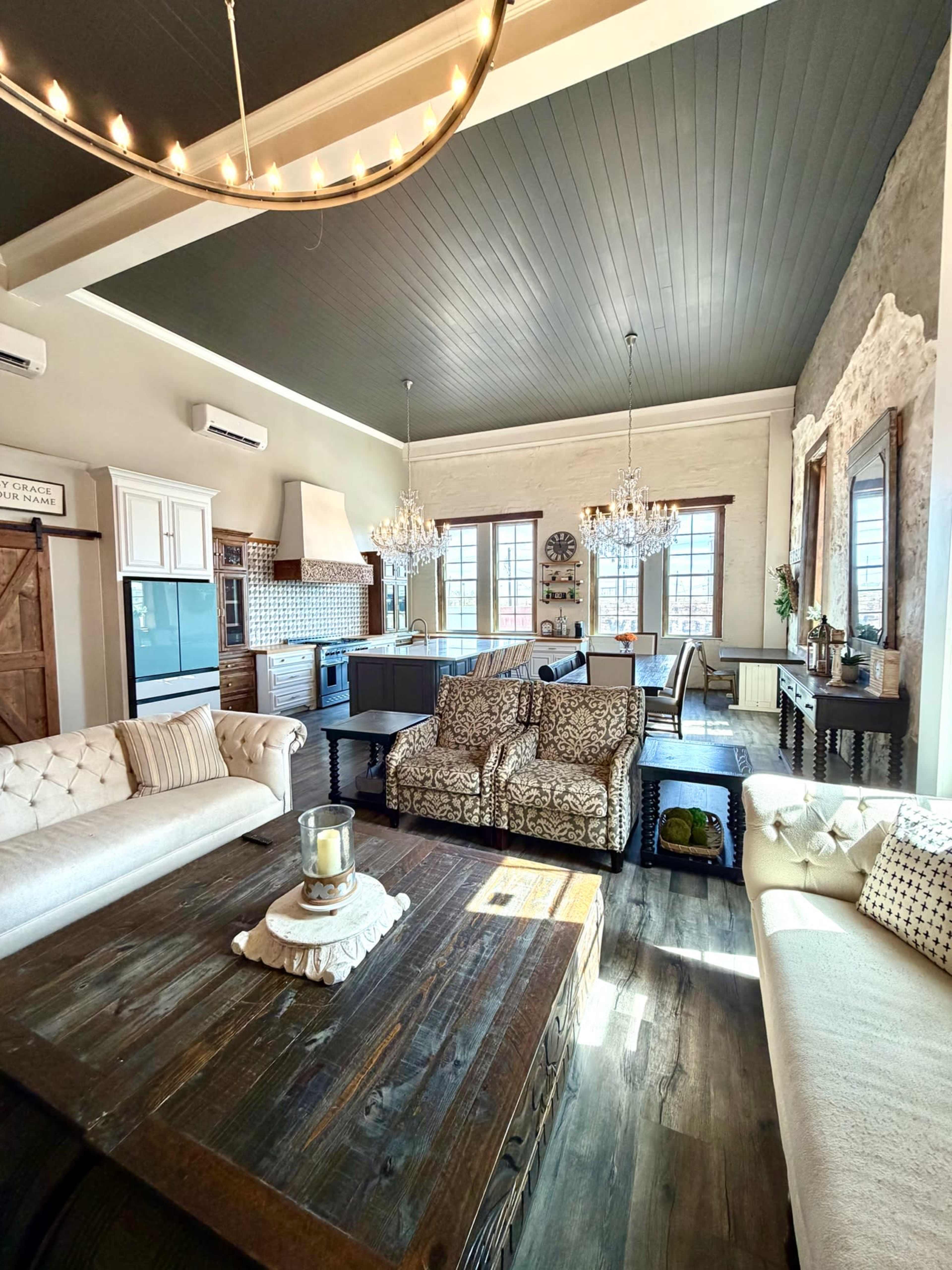 The image shows a spacious living room with a high ceiling, featuring a mix of modern and rustic decor, including a large wooden table, beige sofas, and a kitchen area in the background.