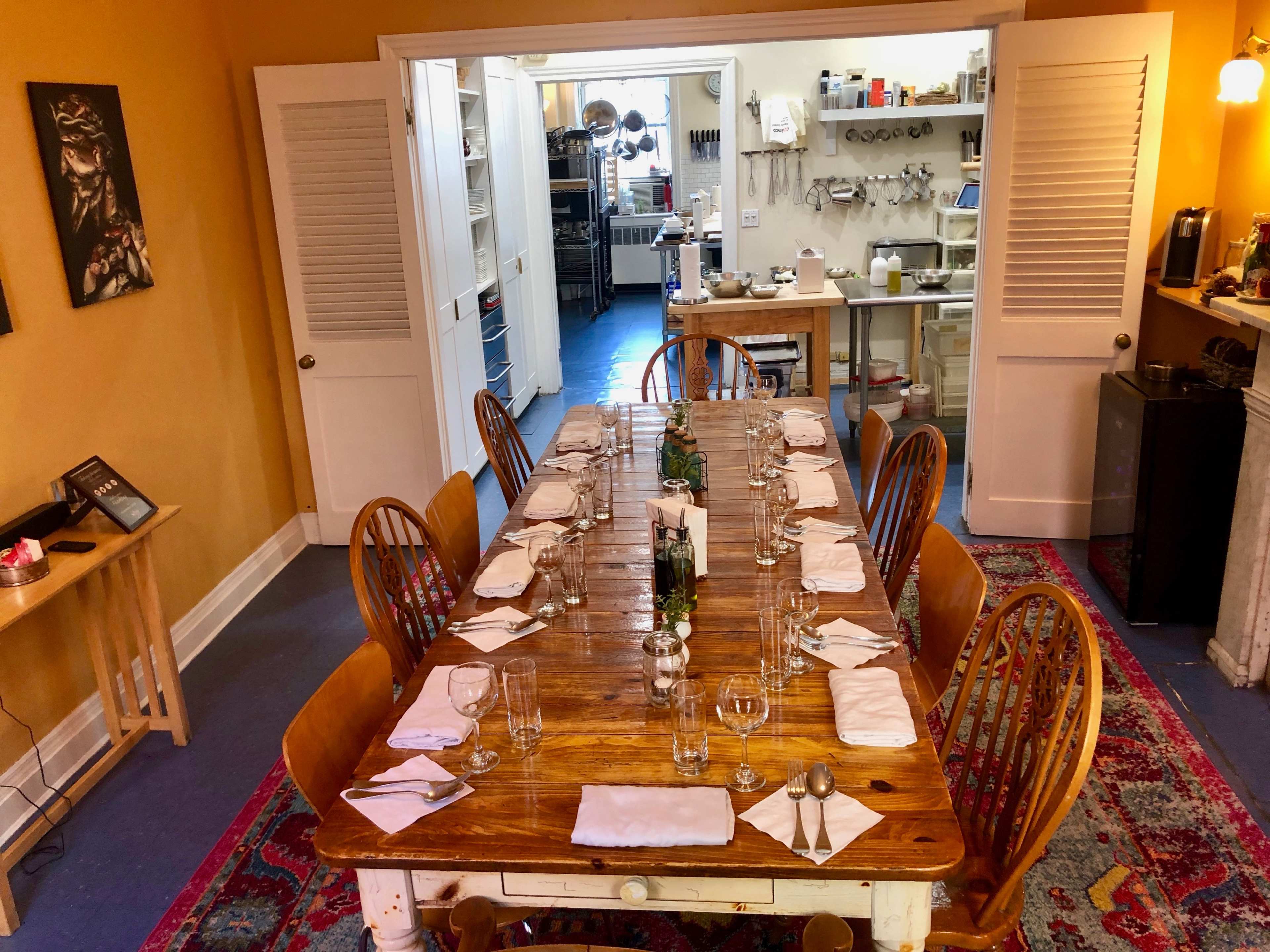 A long wooden dining table is set with glasses and napkins in a brightly lit kitchen space with open doors leading to a kitchen area.