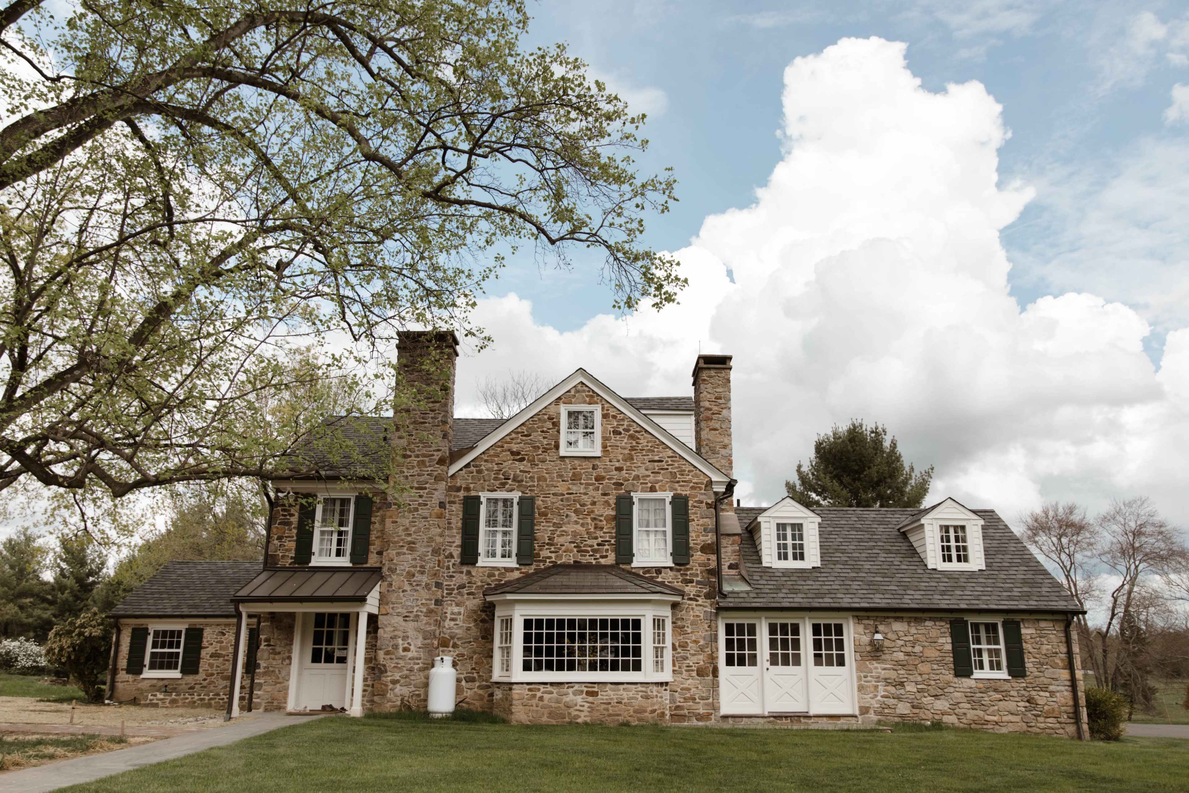 A large stone house with multiple gables and a prominent front porch is set against a backdrop of clouds and trees.