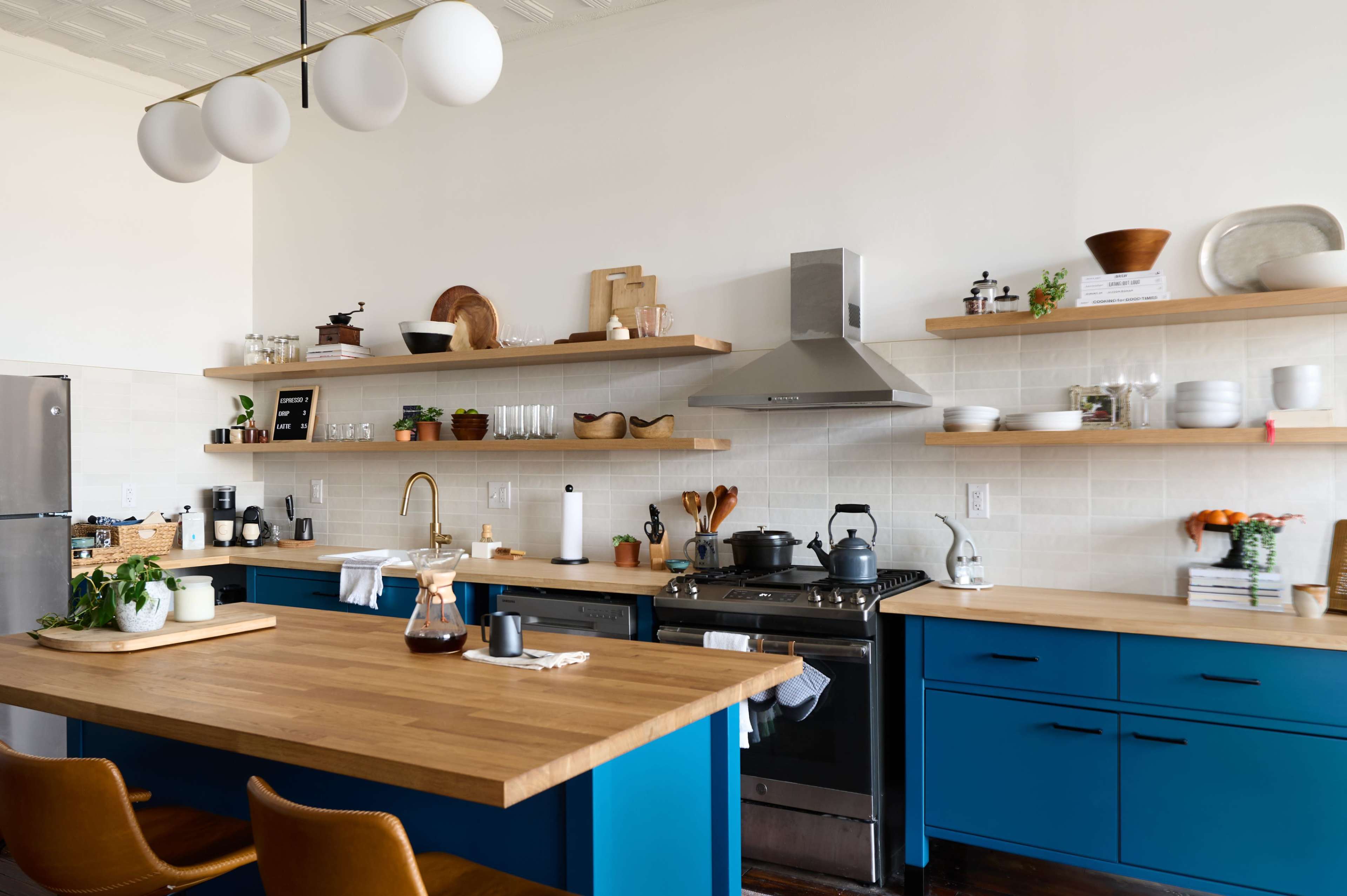 A modern kitchen features a central wooden island with blue cabinetry, open shelving displaying dishes and plants, and stainless steel appliances.