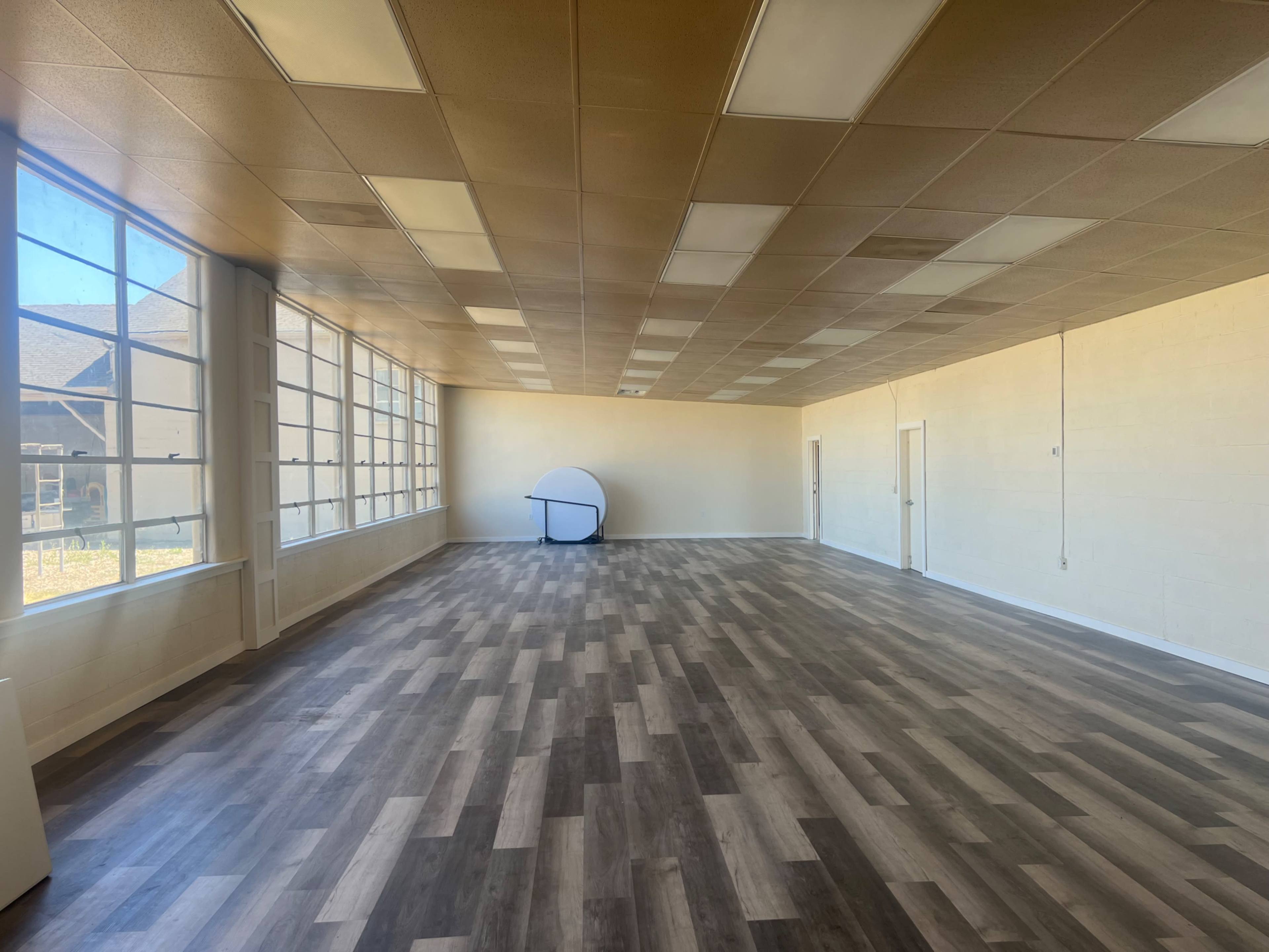 A spacious, empty room with large windows and wooden flooring, featuring a circular table in the center.