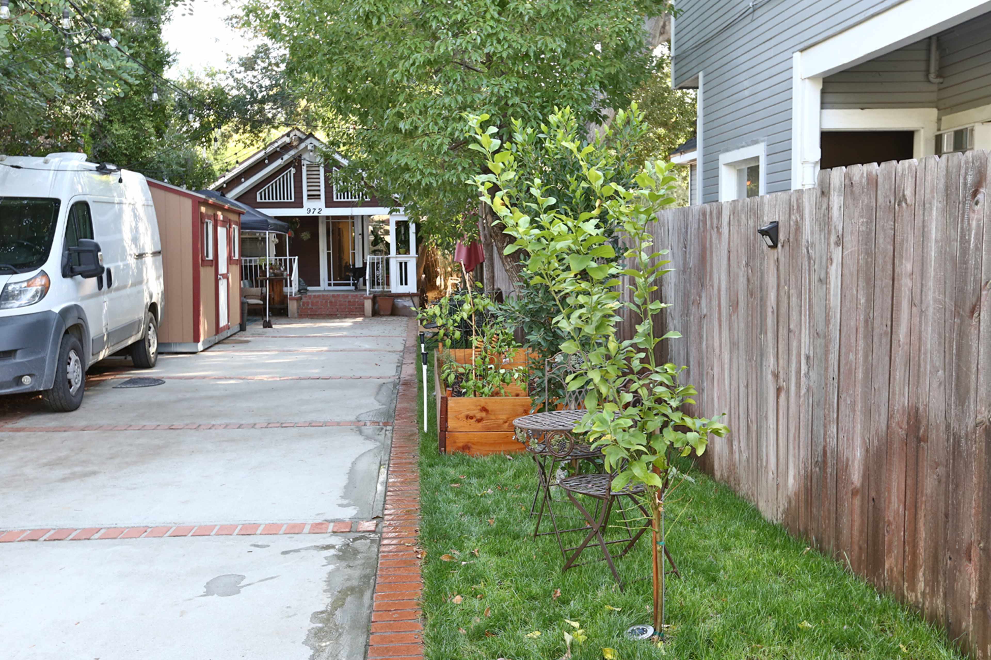 A narrow pathway lined with potted plants and small tables leads to a house set back from a driveway with a van parked nearby.