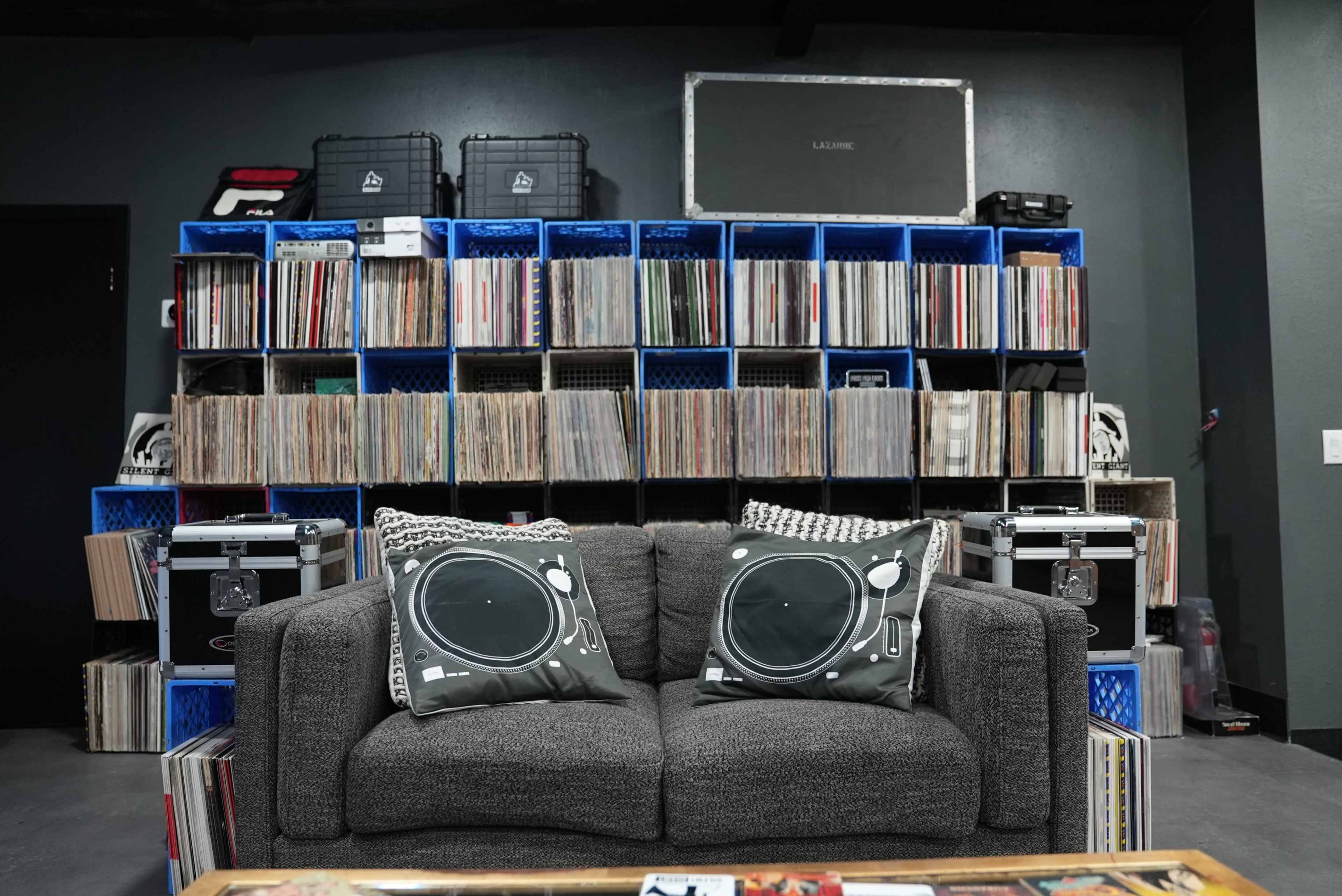The image shows a cozy seating area with a grey couch adorned with turntable-themed pillows, positioned in front of a large shelving unit filled with vinyl records and storage boxes.