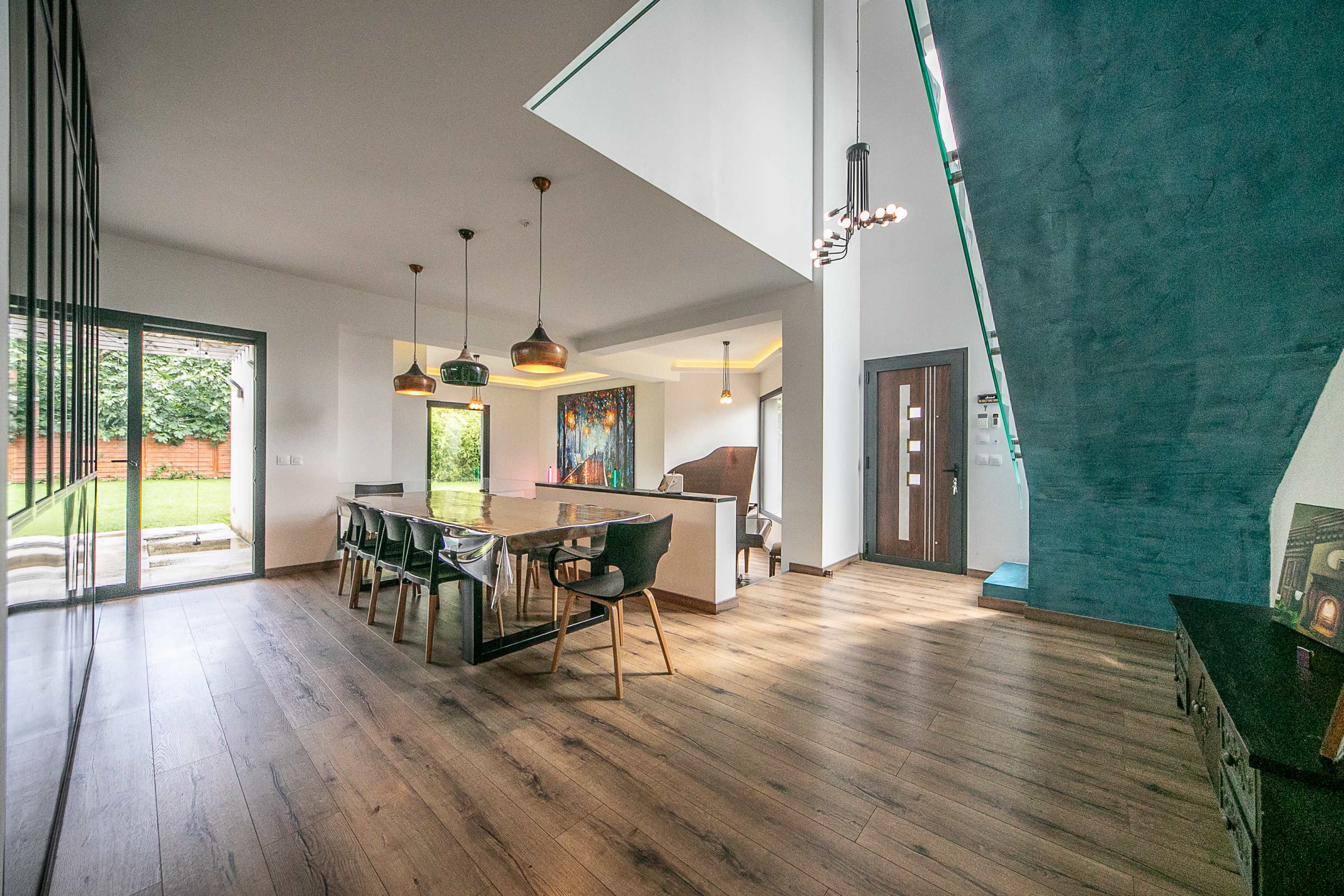 A modern living space featuring a dining area with a large table, chairs, and a kitchen, all connected to a staircase and large windows overlooking a garden.