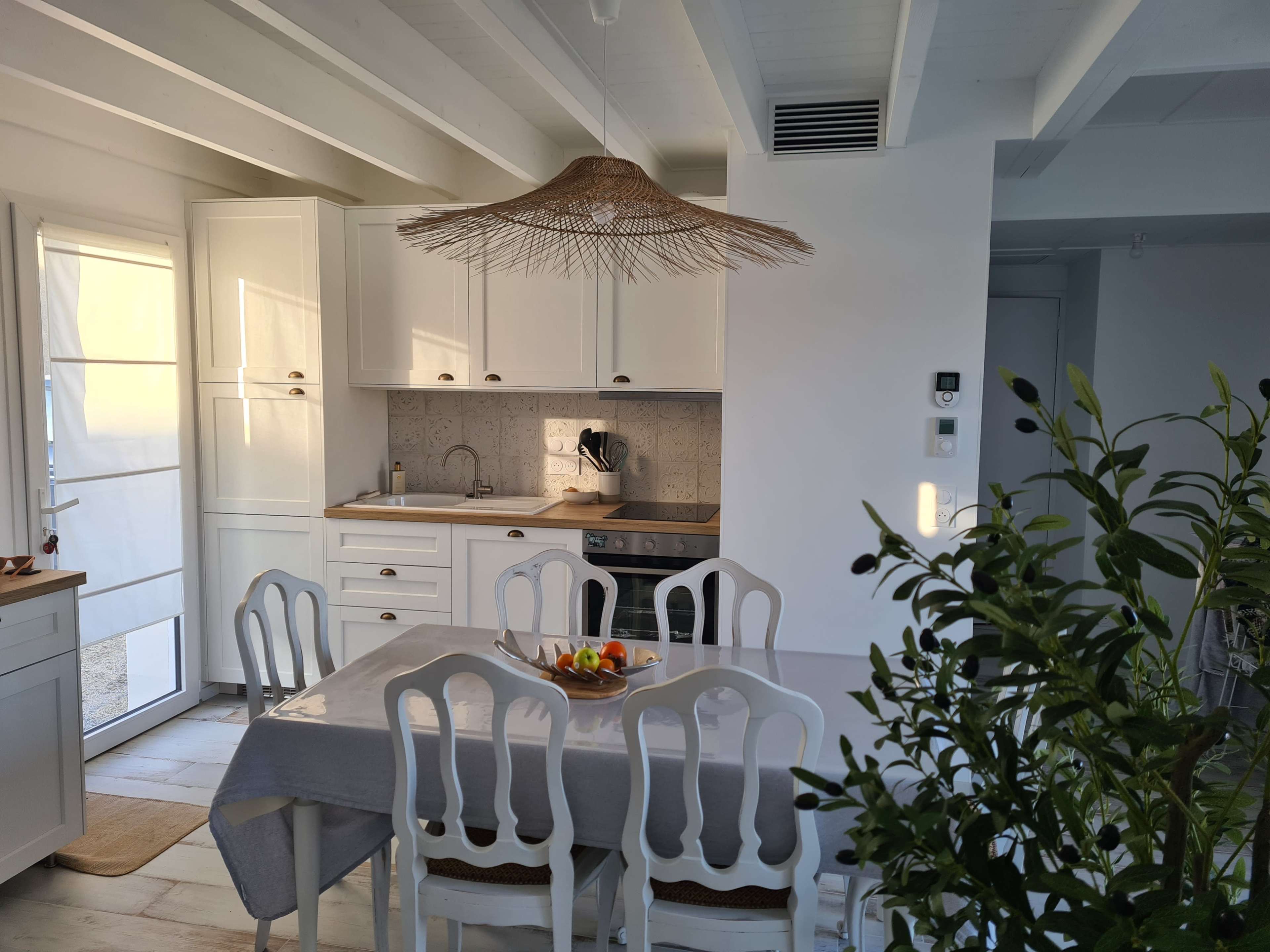 A bright kitchen and dining area features white cabinetry, a wooden table set for meals, and a rattan pendant light.