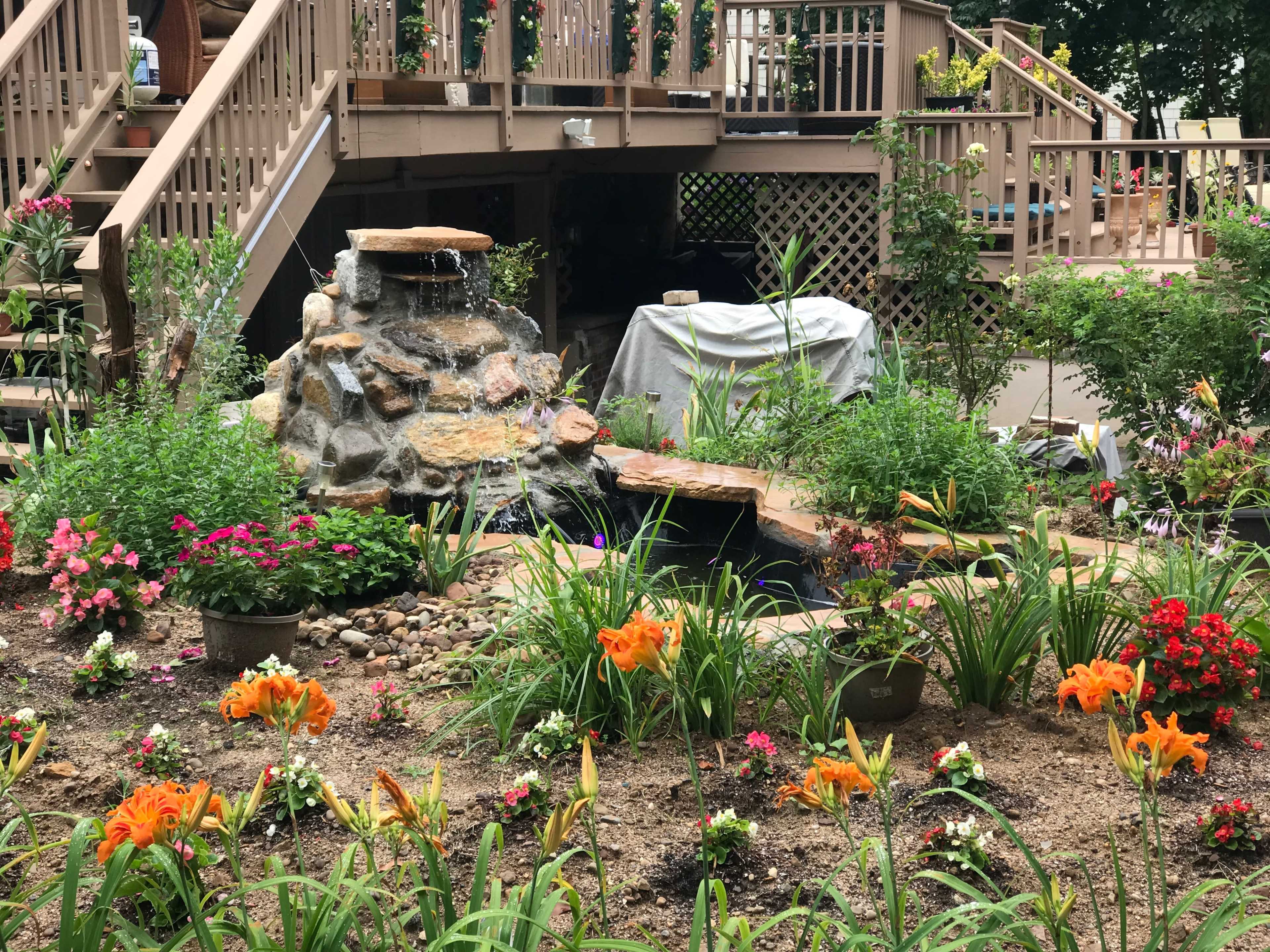 A rock garden with a waterfall is surrounded by colorful flowers and greenery beneath a wooden deck.