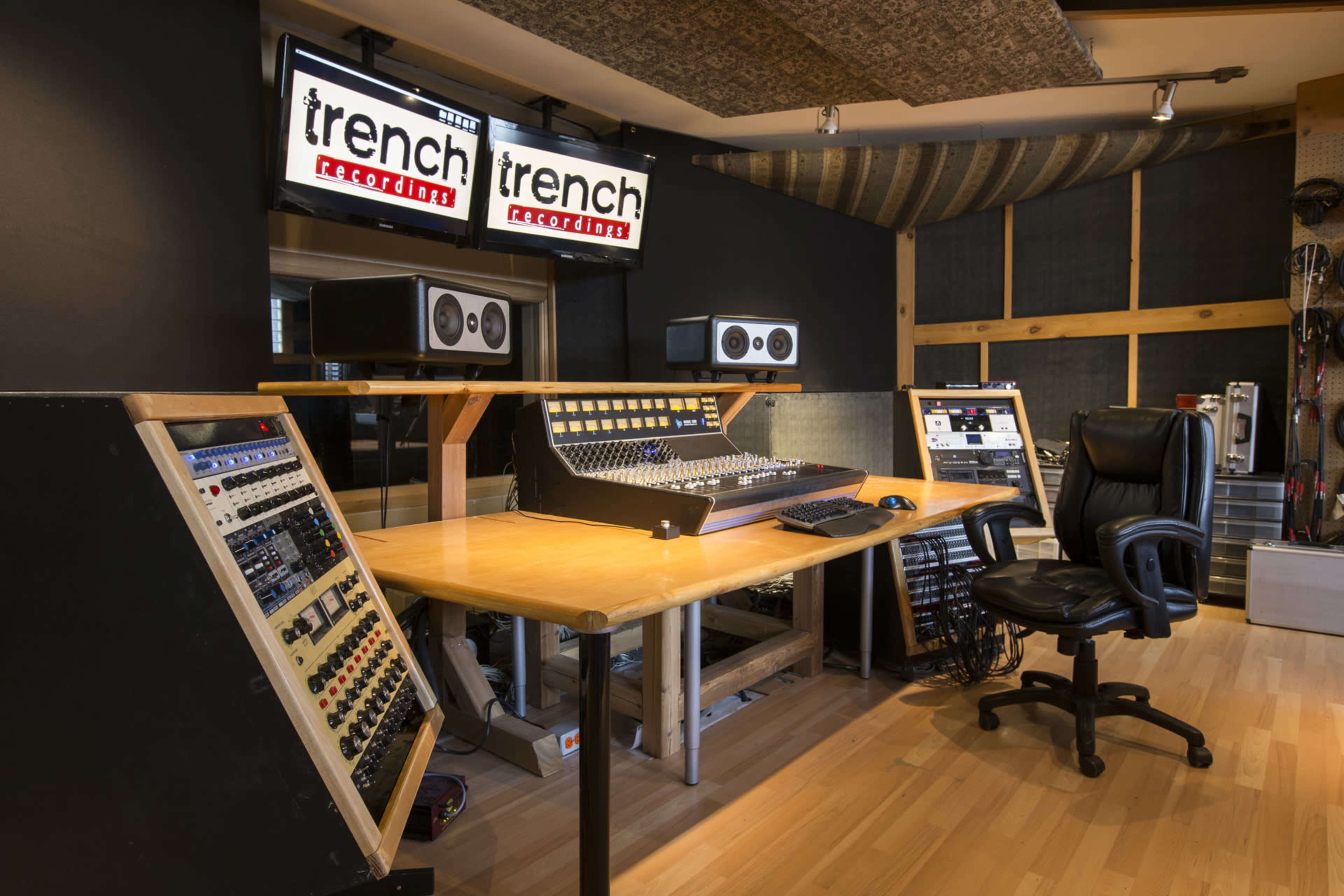 A music recording studio with a large mixing console, multiple sound equipment racks, and two monitors displaying the logo of "Trench Recorders."