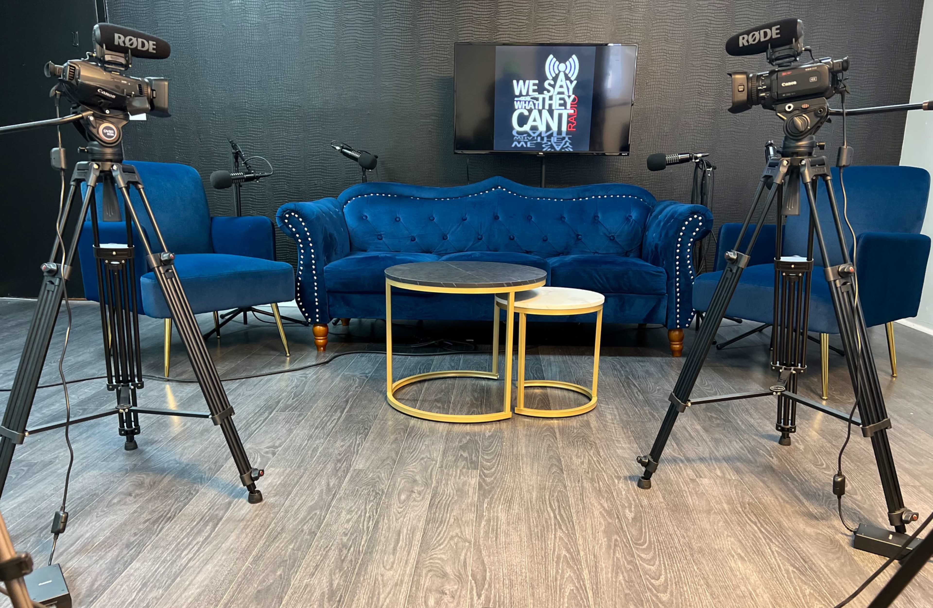 The image shows a video recording setup with two cameras on tripods facing a blue velvet couch and a coffee table in a modern studio environment.