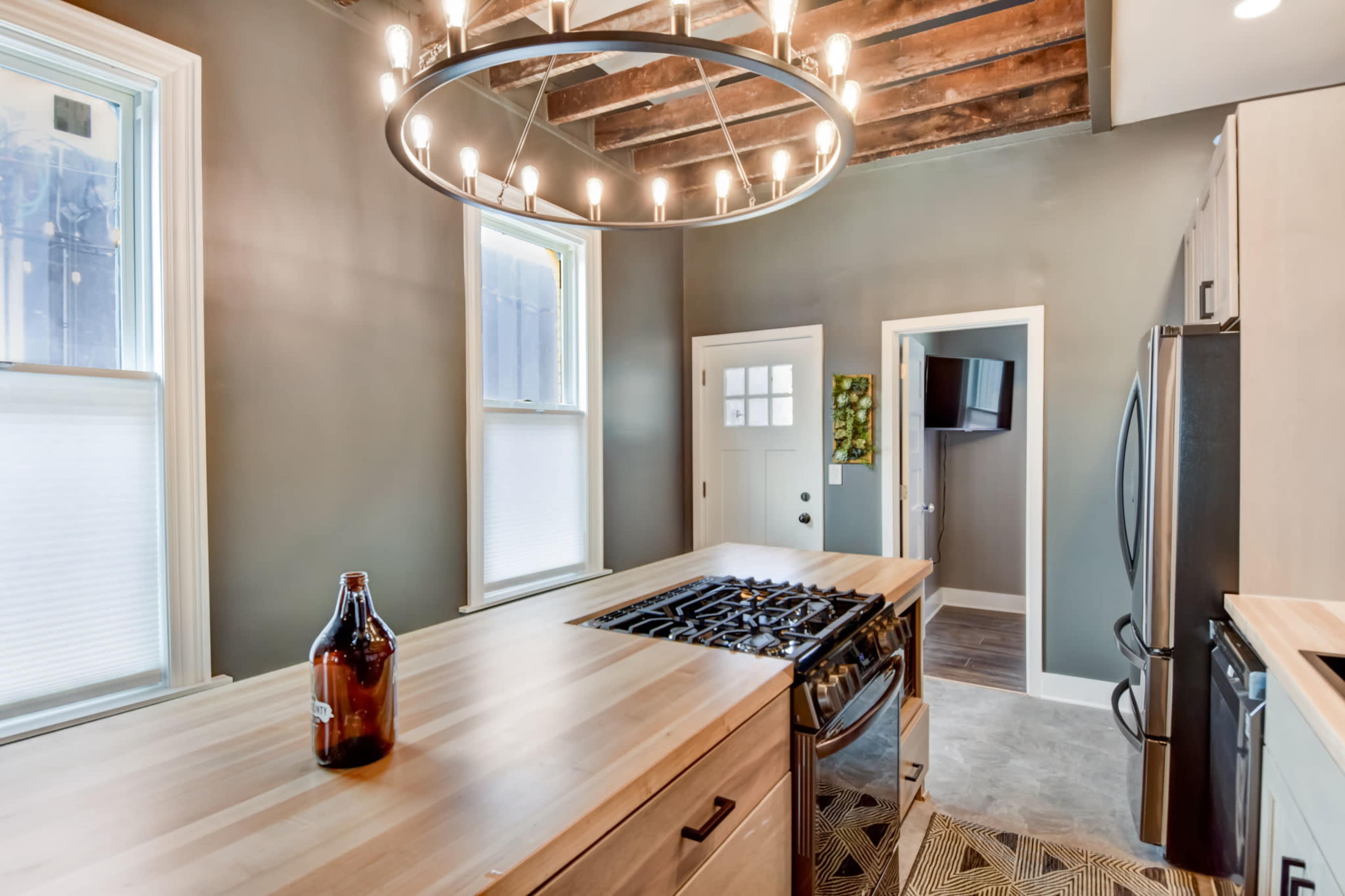 A modern kitchen features a central wooden island with a gas stove and a large circular chandelier above, complemented by exposed wooden beams and gray walls.