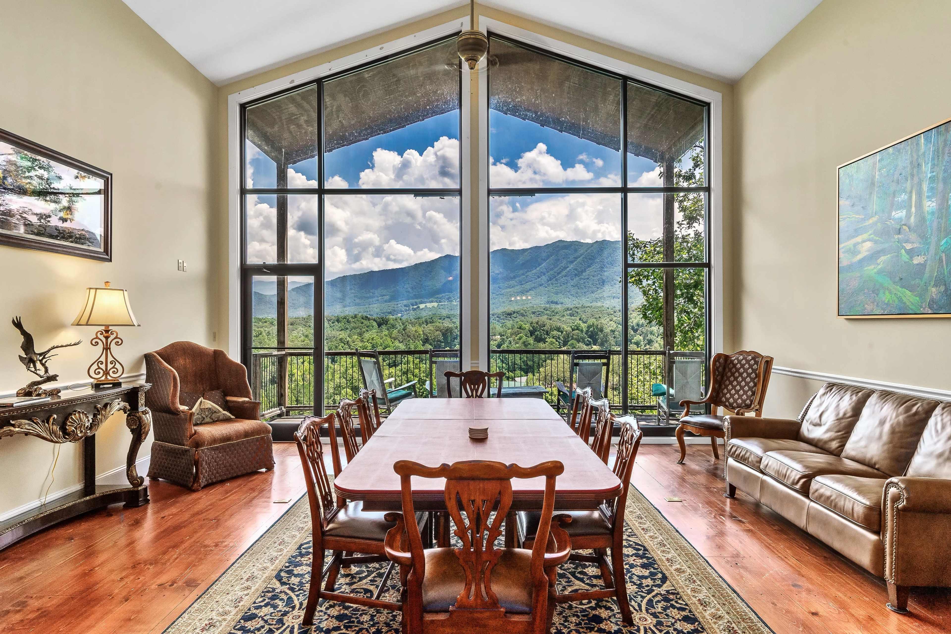 A spacious dining area with a long wooden table is surrounded by large windows that offer a view of the mountains and sky.