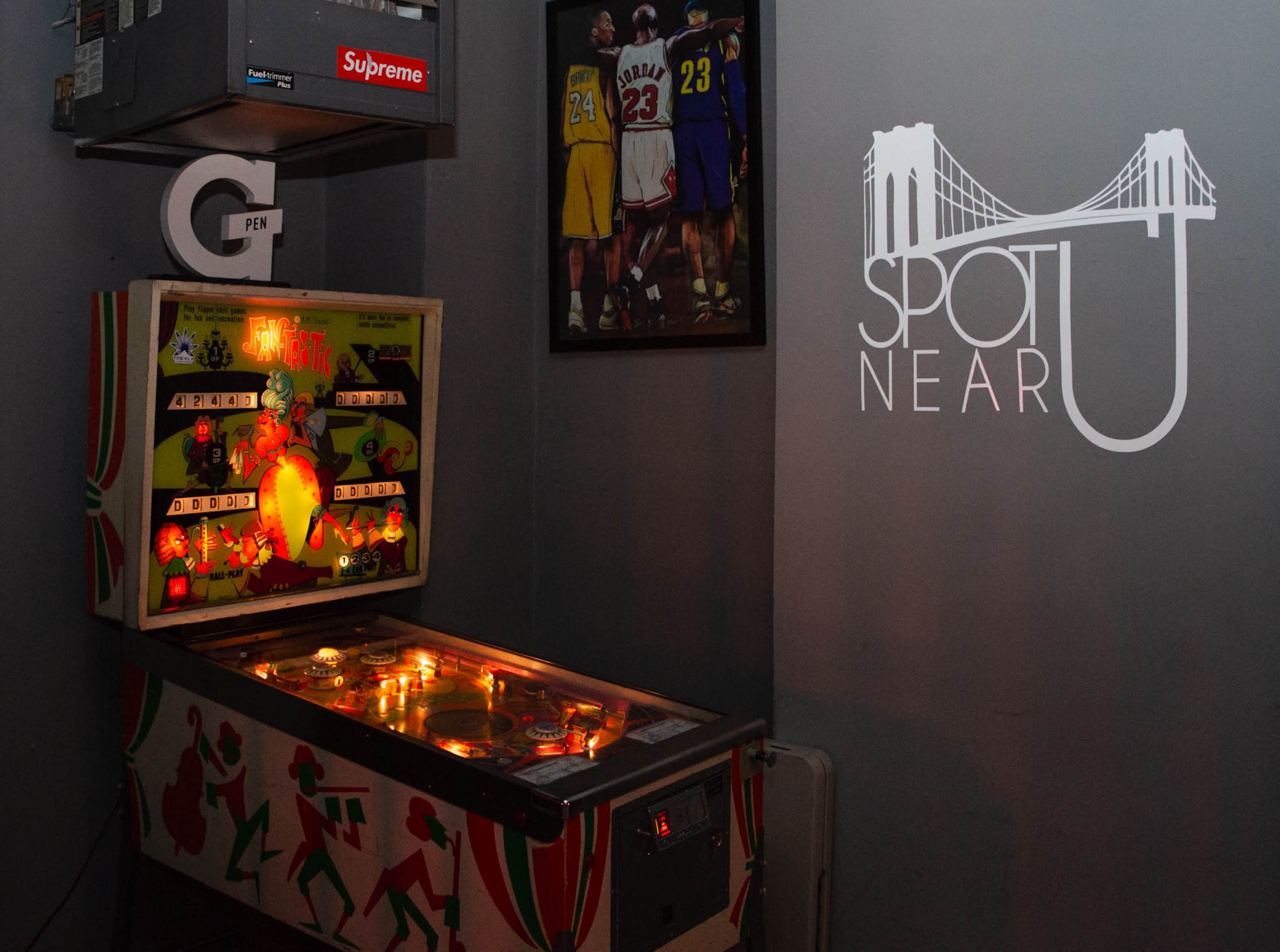 A vintage pinball machine is illuminated in a dimly lit corner, next to a wall featuring a framed image of basketball players and a logo with the text "SPOT NEAR U."