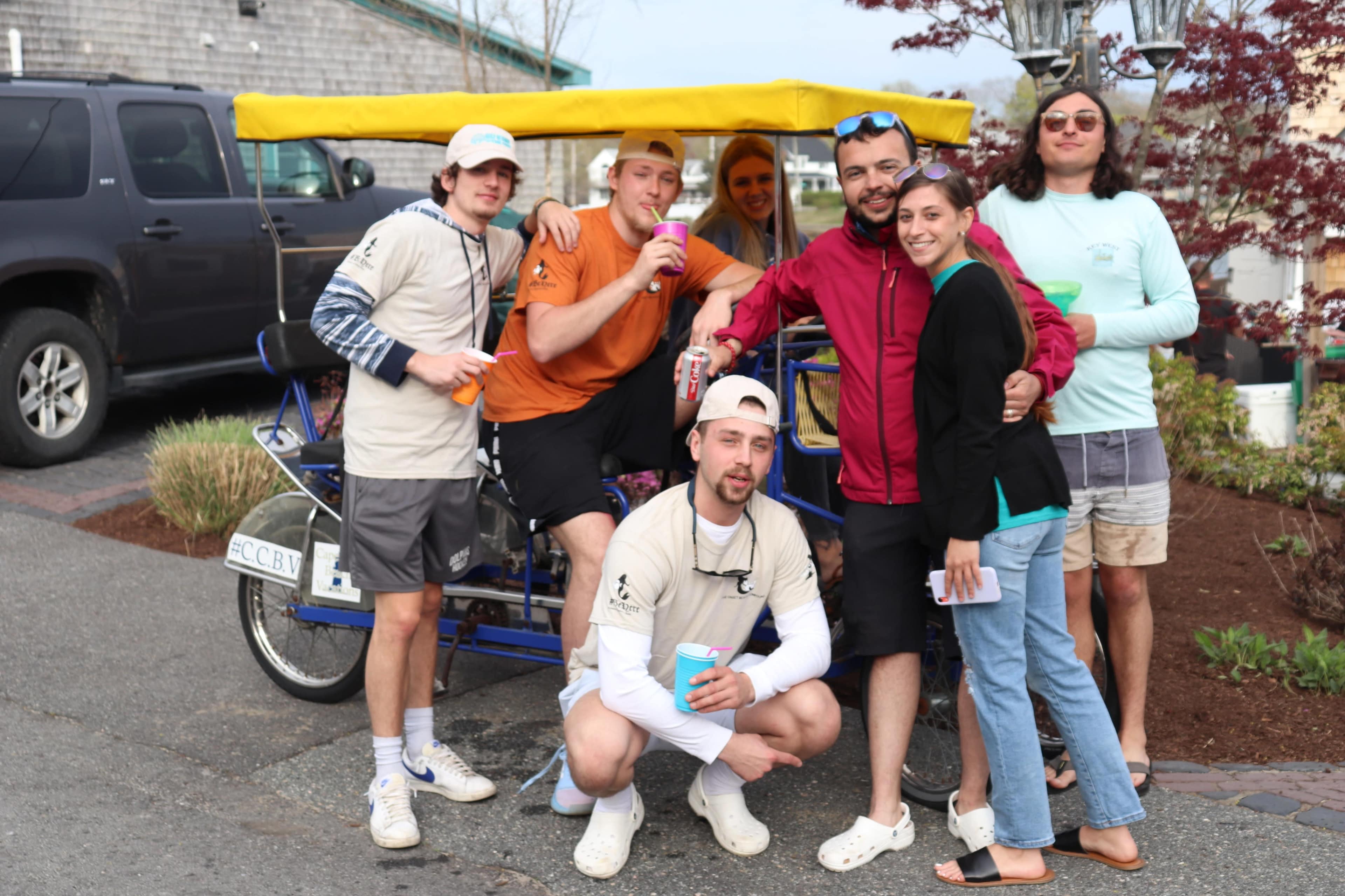 A group of seven young adults poses together near a colorful pedicab in a parking area.