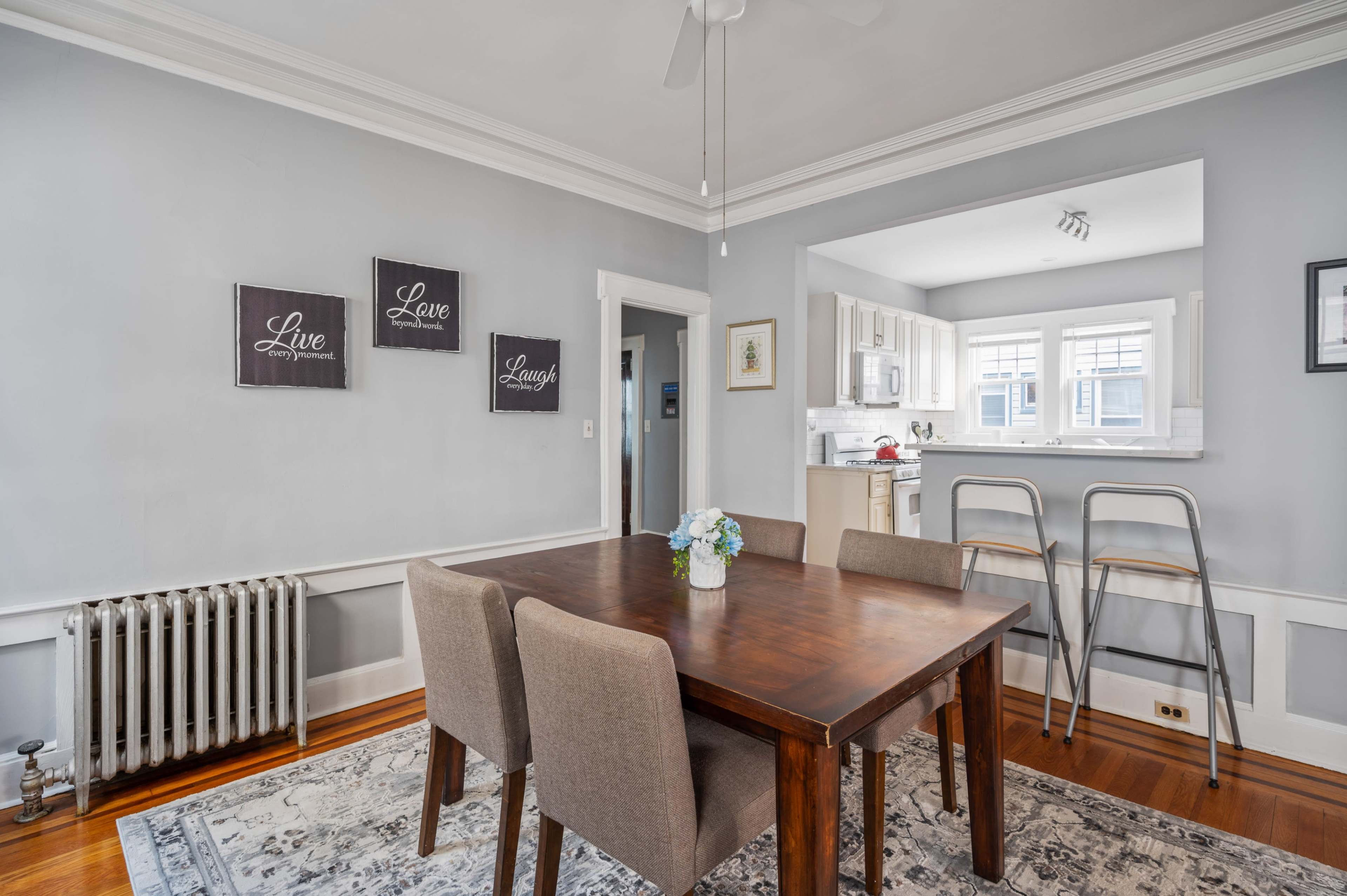 A dining area features a wooden table with four chairs, flanked by a decorative rug and pale gray walls with framed motivational signs.