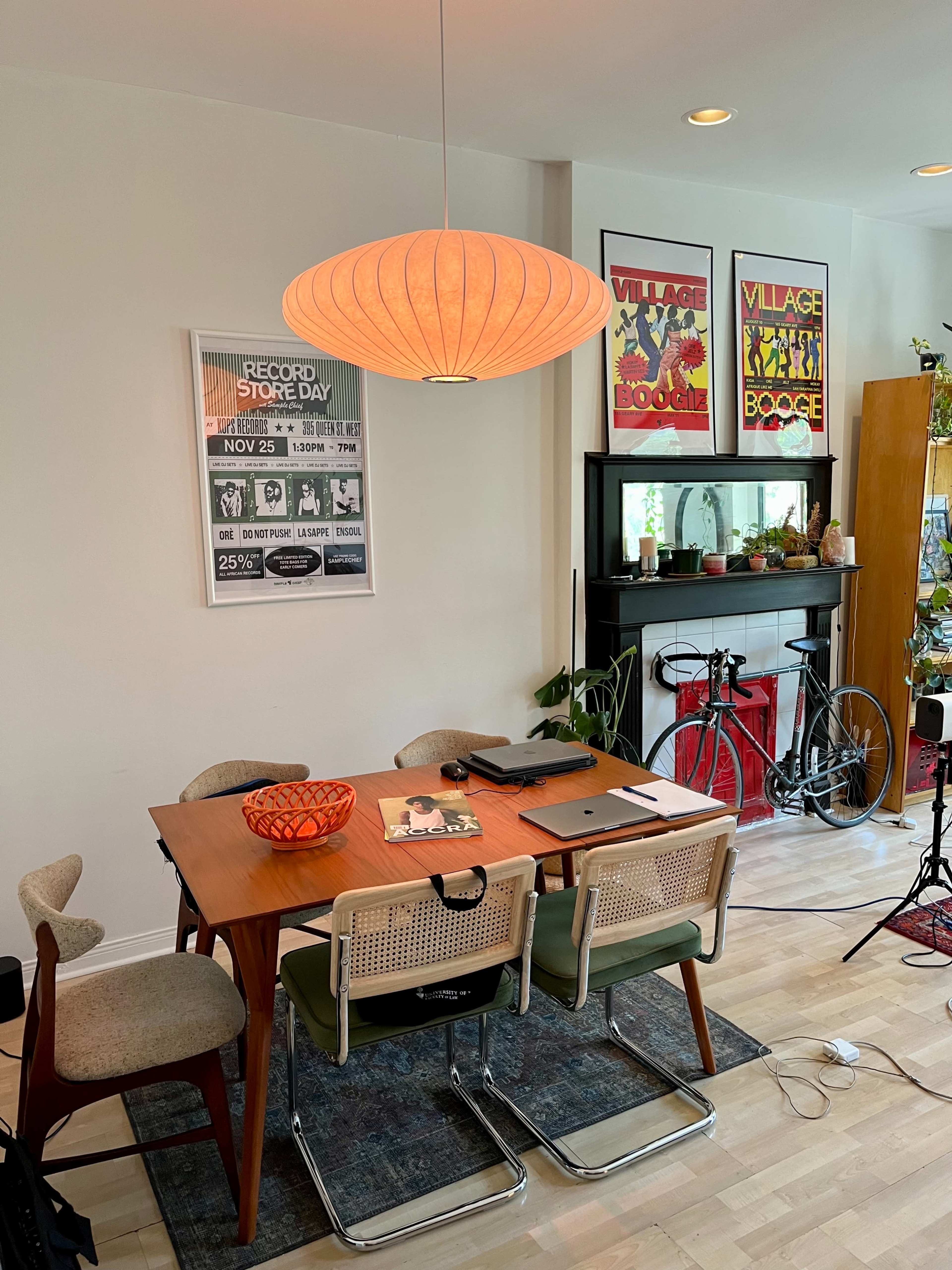 A dining table with four chairs and various items rests in a brightly lit room, featuring posters on the wall and a bike near a glass cabinet.